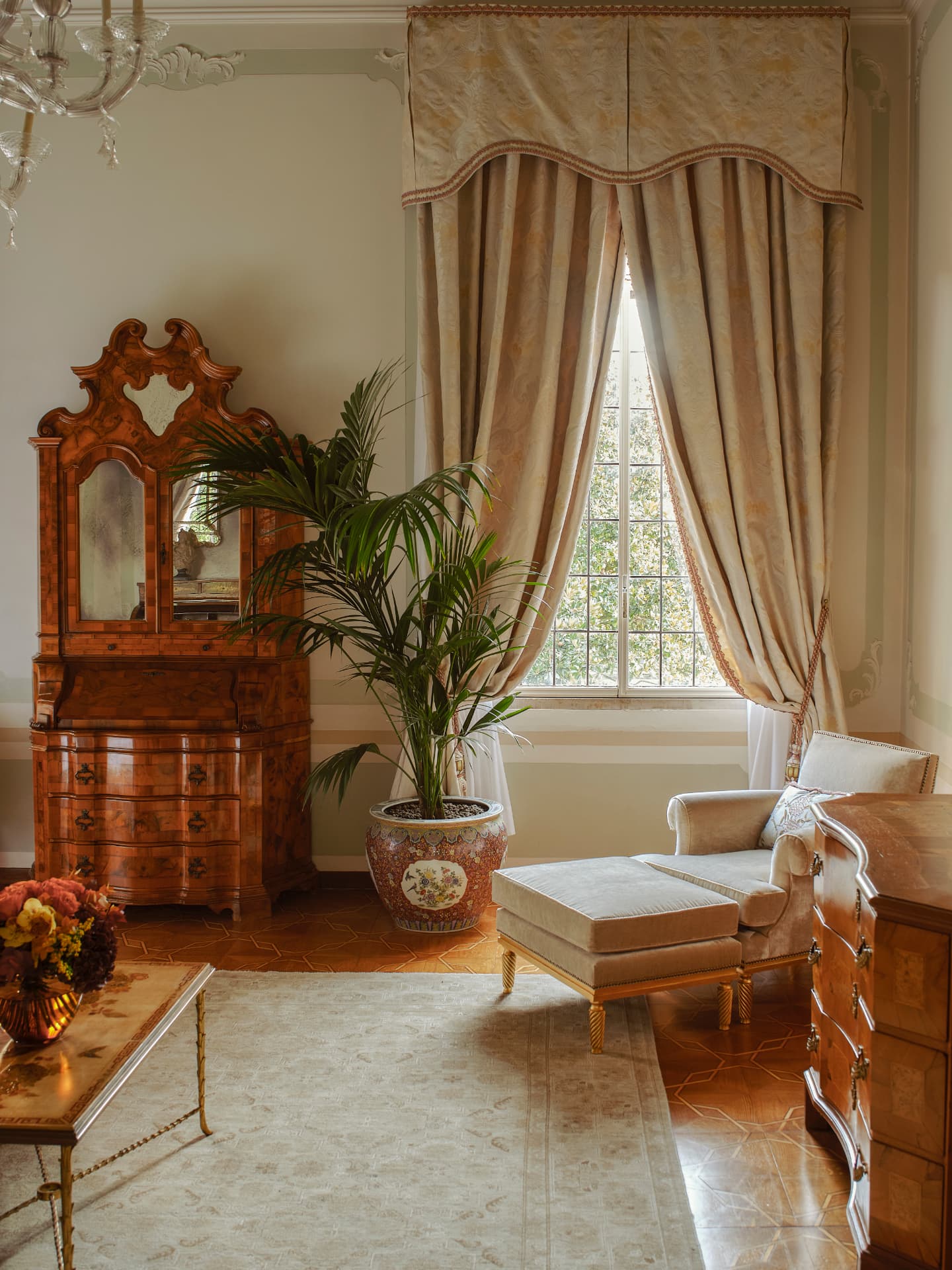 Wohnbereich einer Suite mit venezianischem Sekretär, Kentia-Palme und Sessel am hohen Fenster