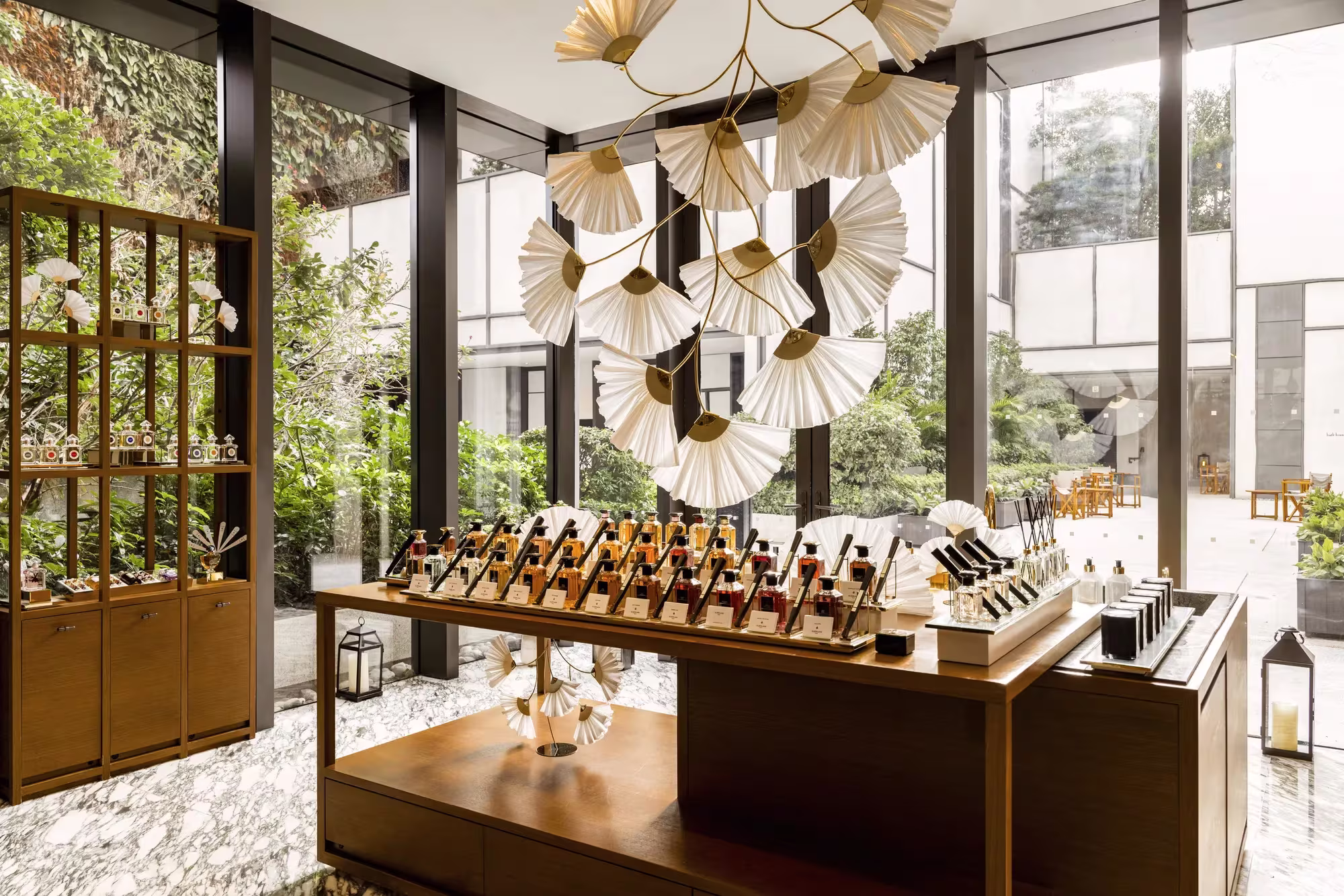 Der Asaya-Bereich im Rosewood Hong Kong mit Fächer-Leuchtenskulptur, Messingregalen voller Pflegeprodukte und Blick in den begrünten Innenhof