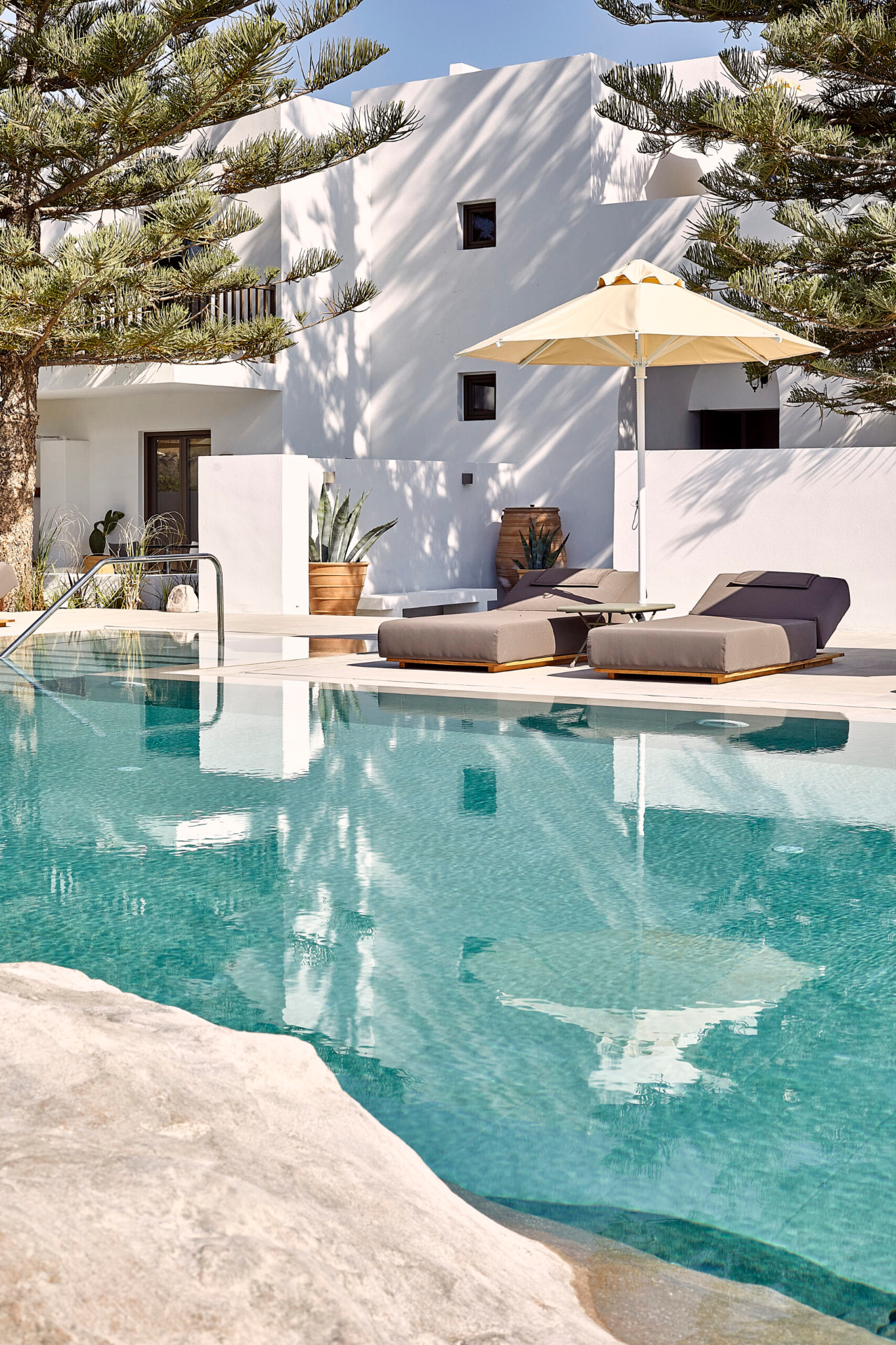 Türkisfarbener Pool des Parīlio mit Liegen, gelbem Sonnenschirm und weissen Bauten hinter einer Araukarie
