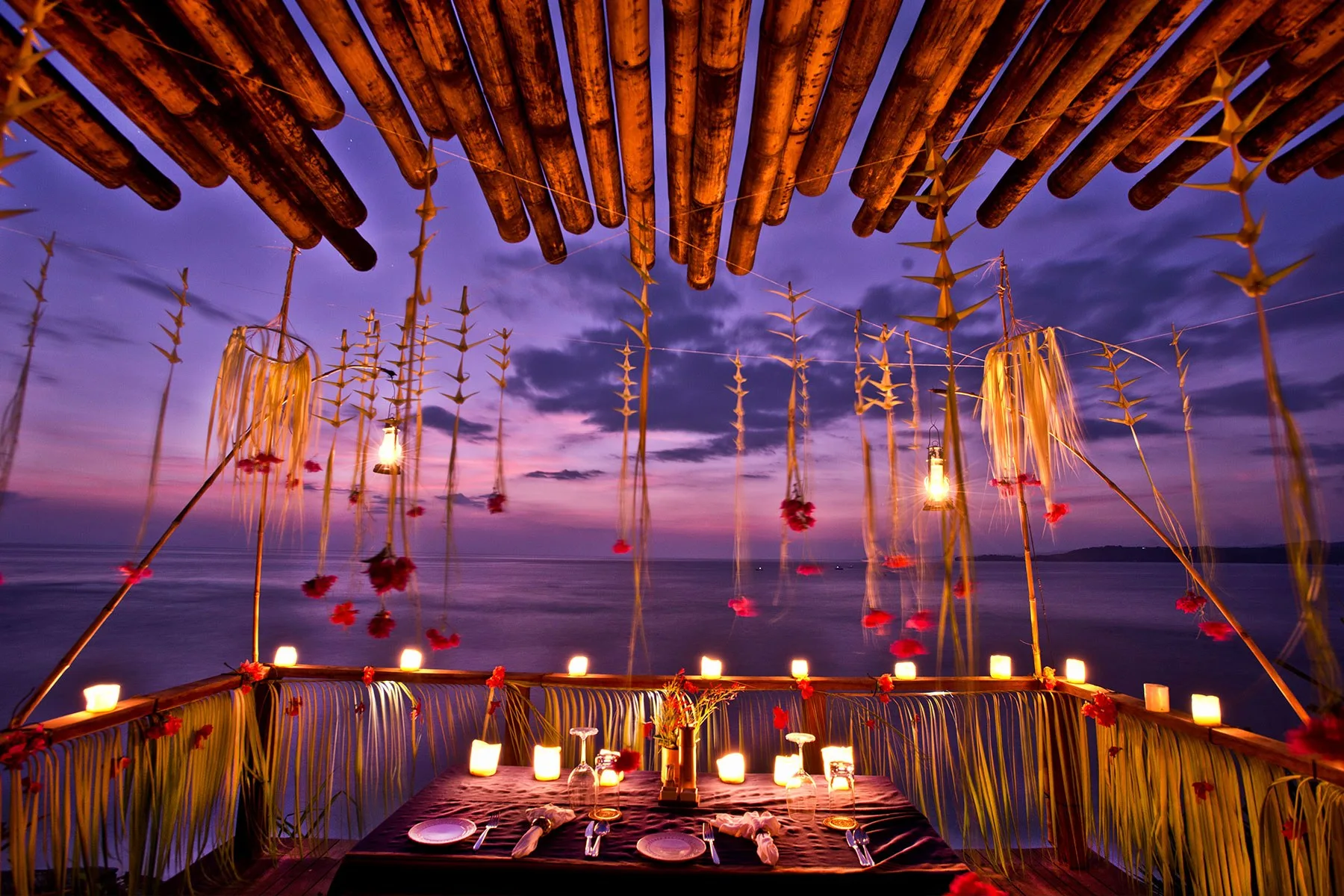 Schlichter offener Tisch auf einer Holzterrasse über dem indonesischen Meer bei Sonnenuntergang, mit gegrilltem Meeresgetier, Gartengrün, tropischen Früchten und Sambal in einer Steinschale