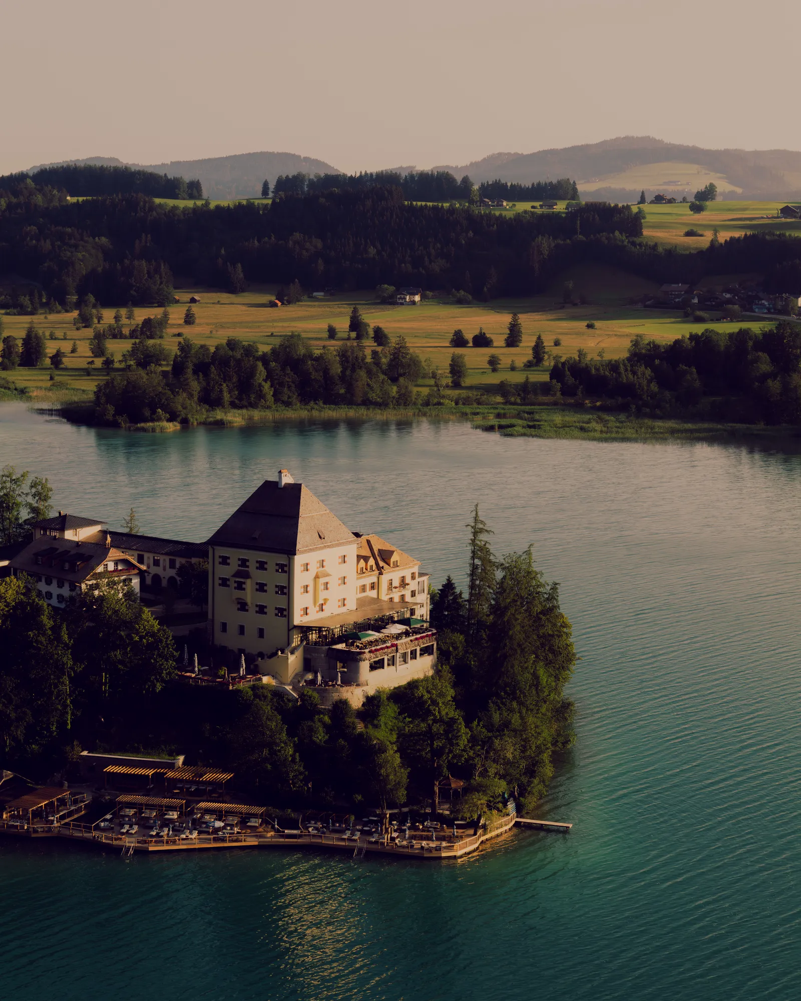 Das Schloss Fuschl auf seiner Halbinsel im türkisfarbenen Fuschlsee, dahinter Wiesen und bewaldete Hügel im Abendlicht