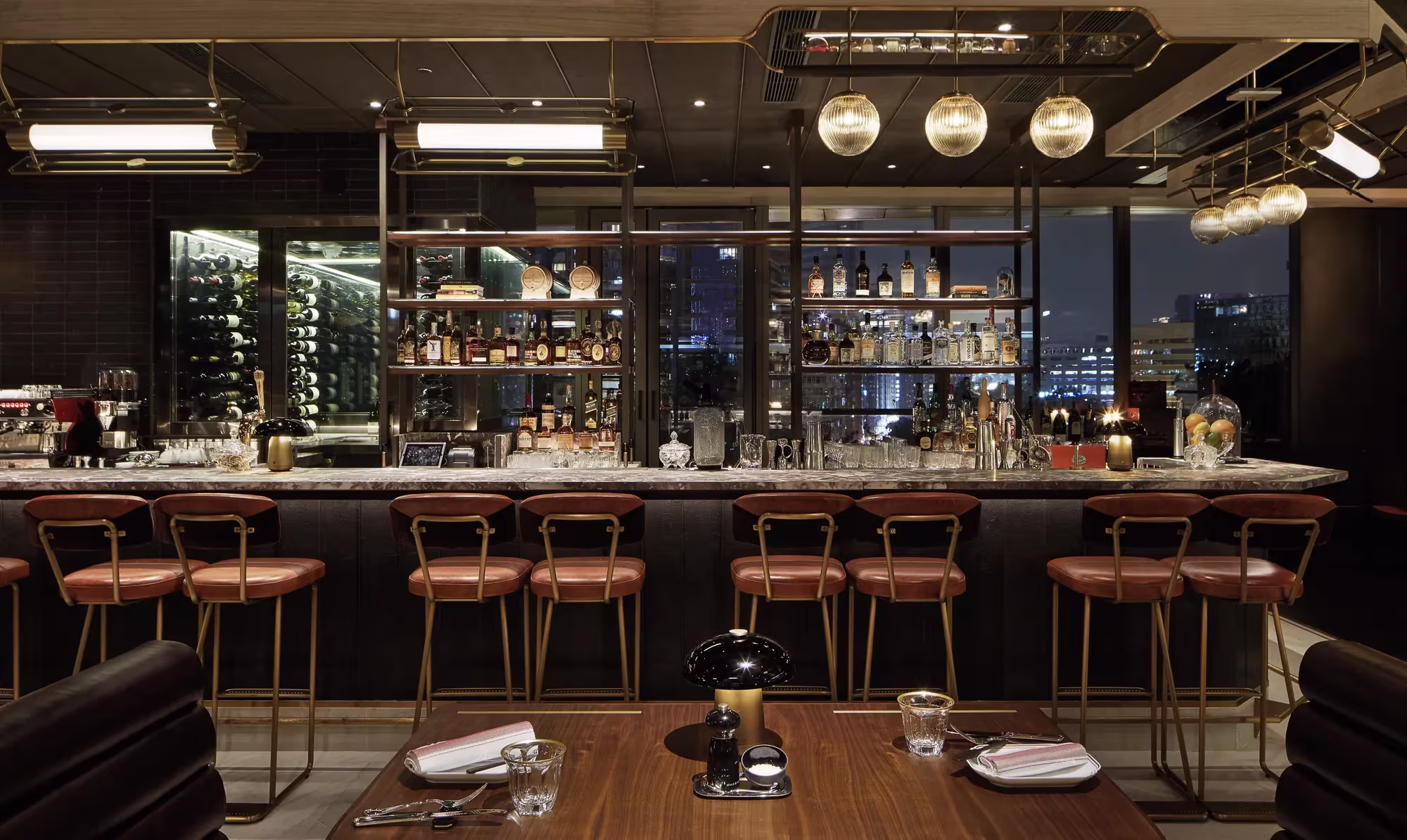 Die Bar des Restaurants Henry im Rosewood Hong Kong mit langem Tresen, Lederhockern und der nächtlichen Skyline hinter den Fenstern