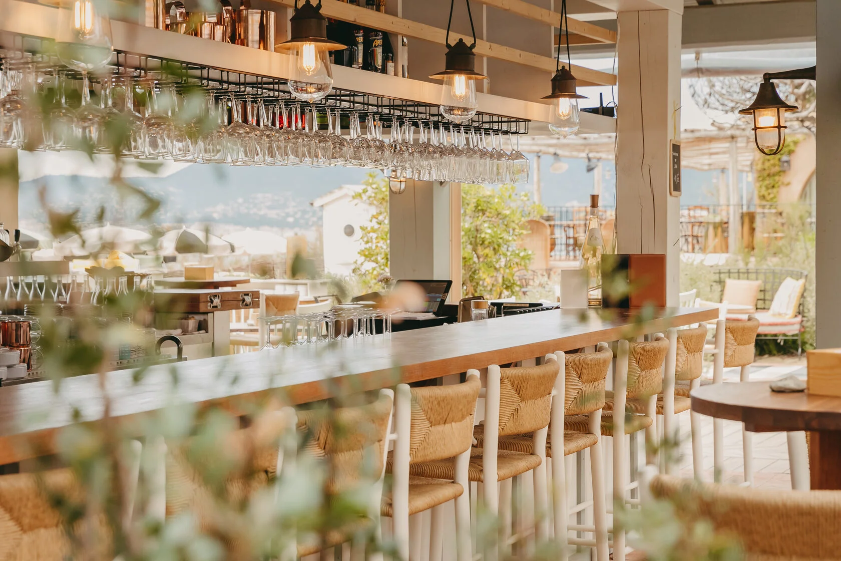 Bar mit Holztresen, Korbhockern und Gläserreihen, dahinter die Bucht im Gegenlicht