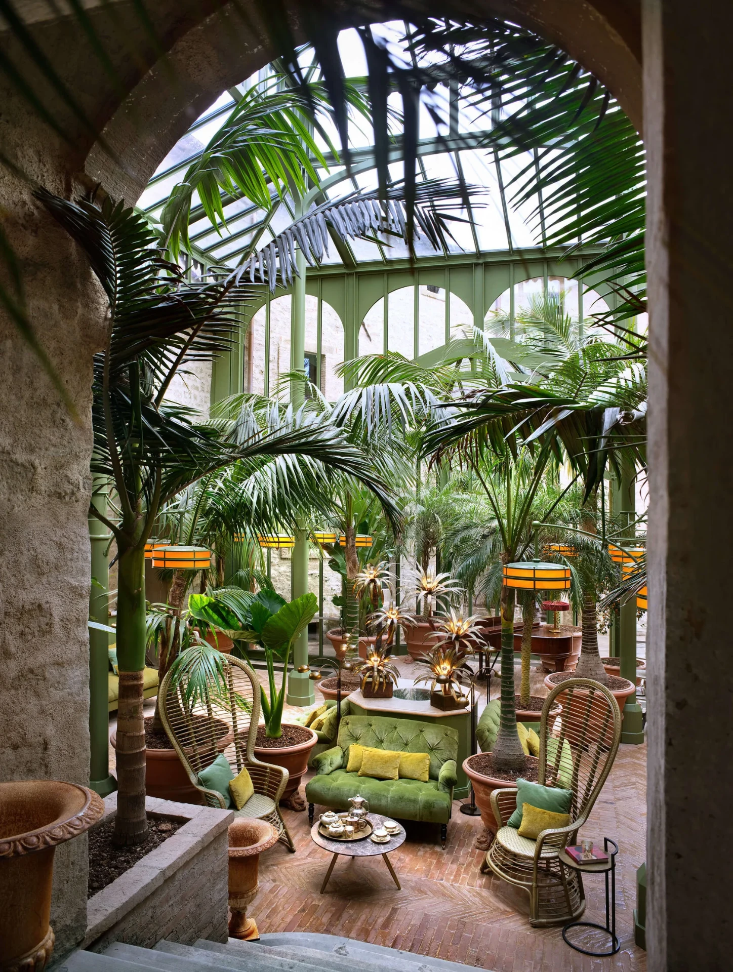 Palm Court mit Palmen unter Glasdach, grünem Samtsofa und Korbsesseln