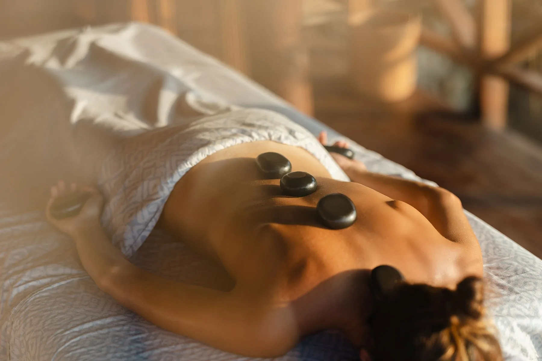 Offener Spa-Pavillon auf einer Klippe in Sumba mit einer schlichten Massageliege unter einem Strohdach, weißer Leinenwäsche im Wind der Meeresbrise und dem Indischen Ozean weit unten