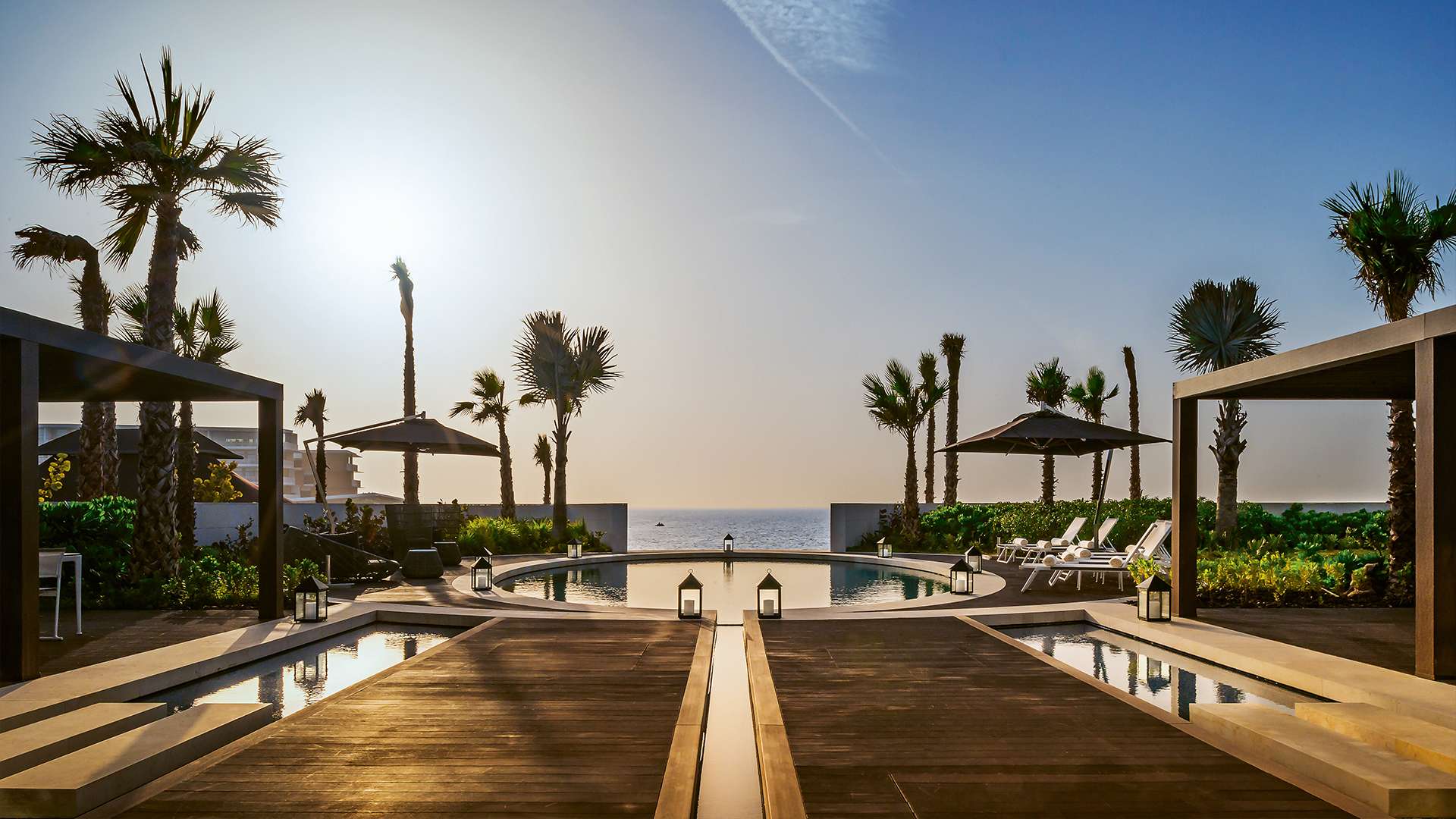 Der runde Strandpool zwischen Palmen und Pavillons, dahinter das offene Meer im Abendlicht