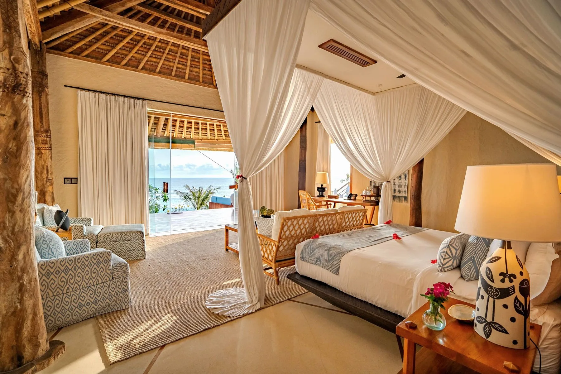Offenes indonesisches Villenschlafzimmer mit Teak-Himmelbett, weißer Bettwäsche, Strohdach und fehlender vierter Wand, mit Blick auf einen privaten Pool und den Indischen Ozean bei Sonnenaufgang