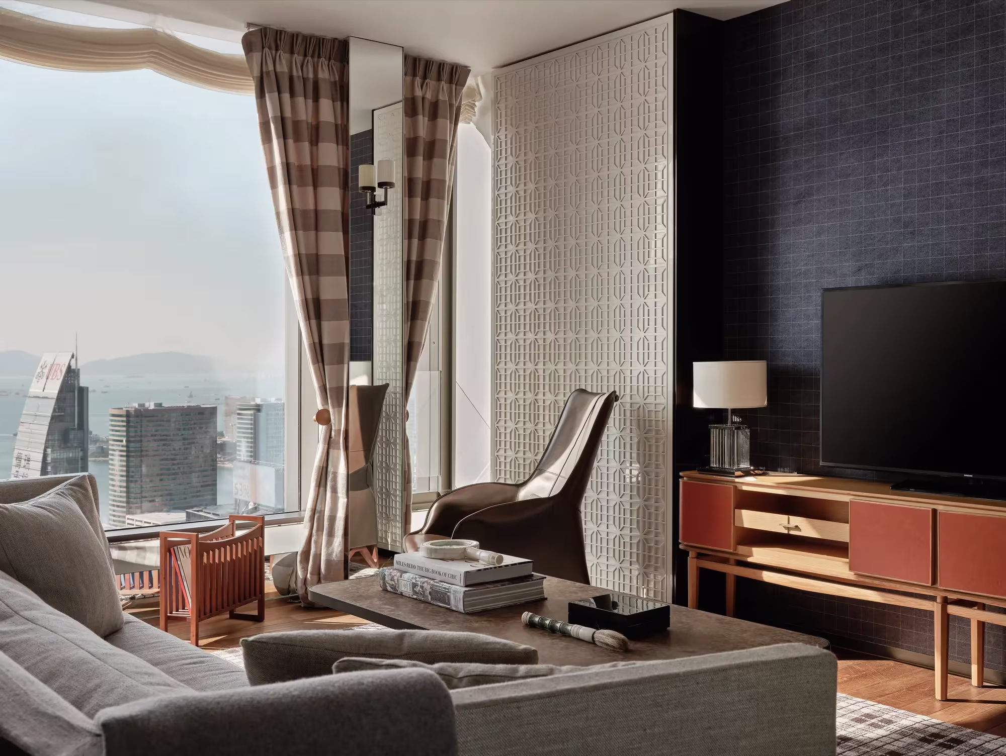 Wohnbereich einer Deluxe Harbour Suite im Rosewood Hong Kong mit grauem Sofa, karierten Vorhängen und Blick über die Skyline