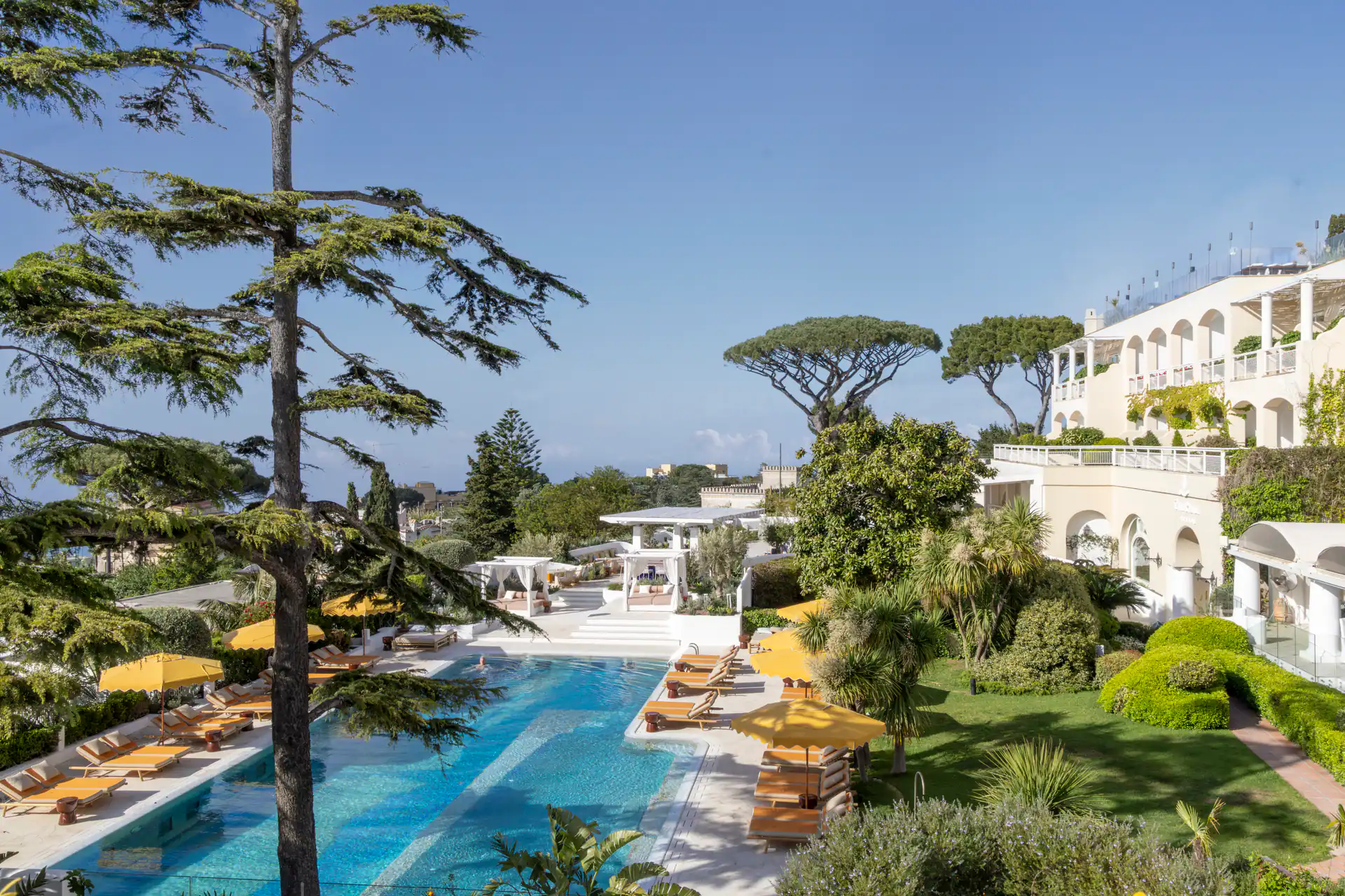 Pool mit gelben Schirmen zwischen Pinien, dahinter das weiße Haus in Anacapri