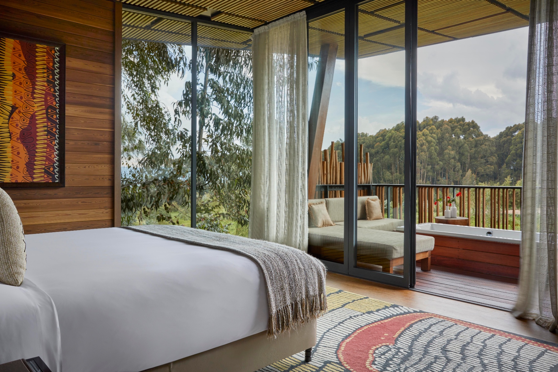 Schlafzimmer aus Holz und Vulkanstein in einer ruandischen Waldlodge vor Sonnenaufgang mit brennendem Kamin, weißem Leinen, Wanderstiefeln an der Tür und nebligem Eukalyptus hinter dem Glas