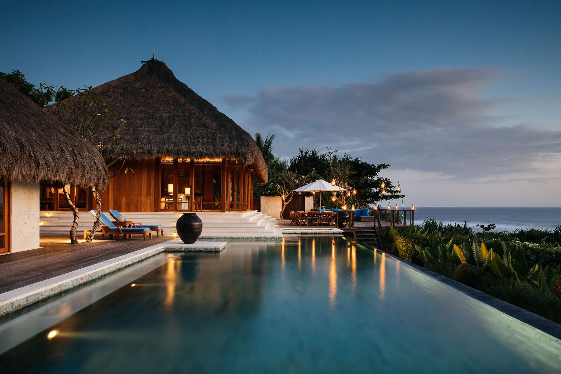 Private Holzterrasse einer strohgedeckten Villa auf Sumba am späten Nachmittag mit einem kleinen Infinity-Pool, der zu einer wilden Bucht hin ausläuft, im goldenen Sonnenuntergangslicht