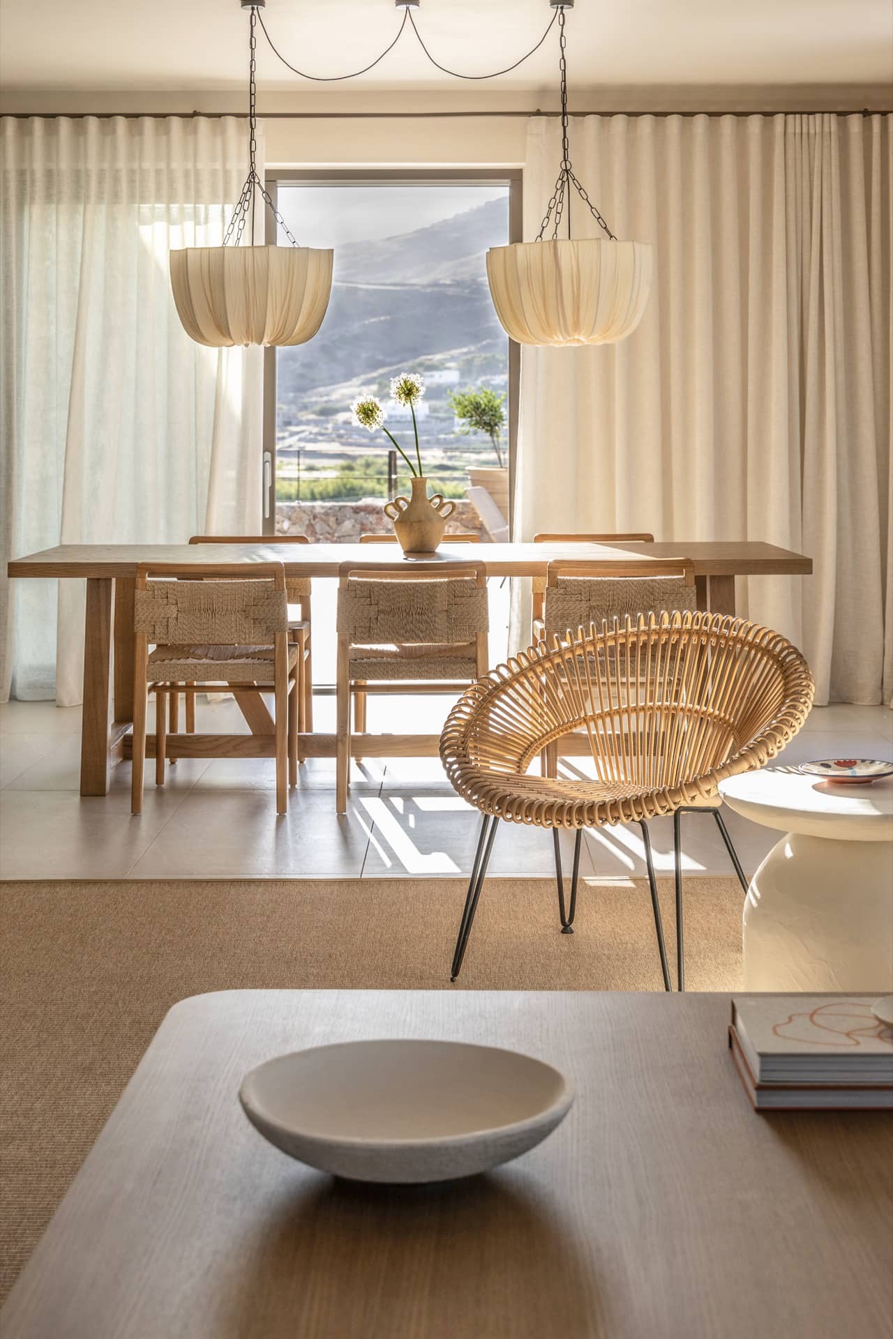 Wohnbereich einer Villa im Parīlio mit Rattansessel, Leinenvorhängen und Esstisch aus heller Eiche im Nachmittagslicht