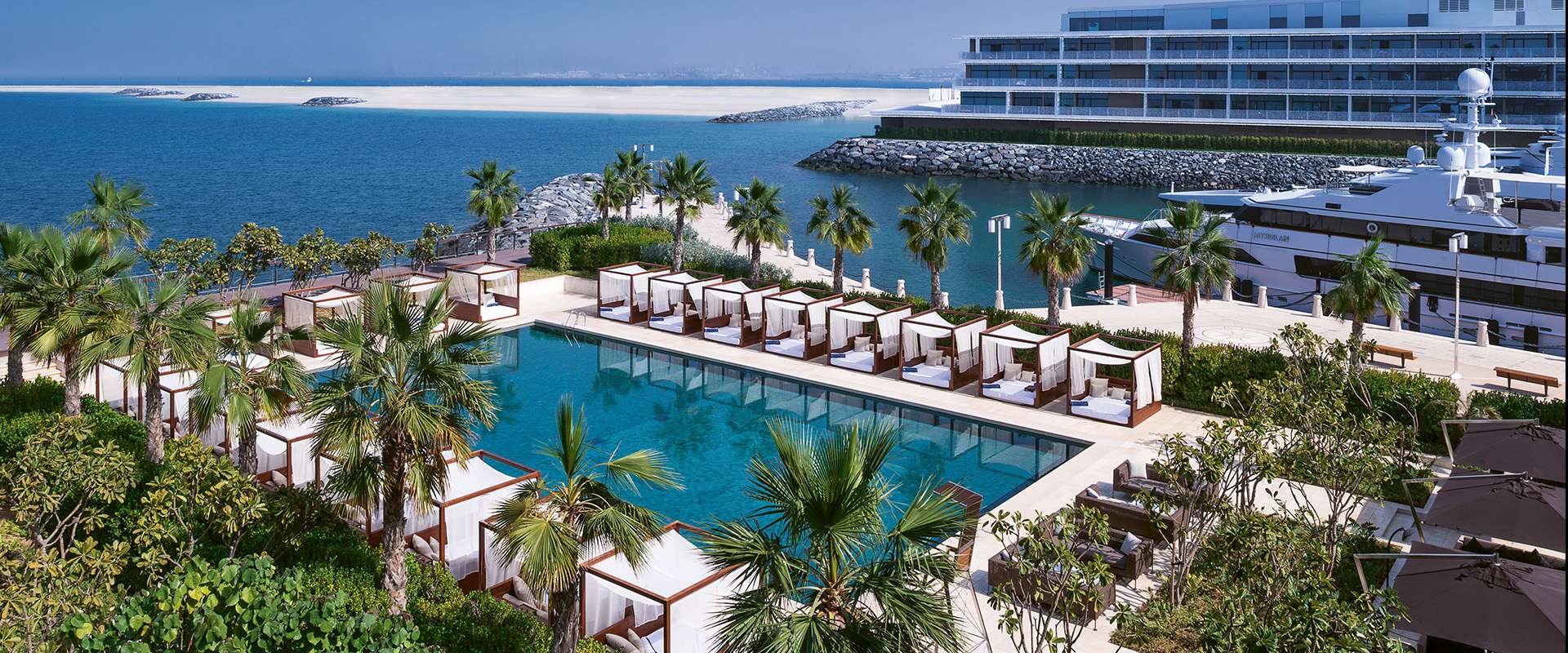 Pool mit weissen Cabanas zwischen Palmen, dahinter der Yachthafen des Resorts