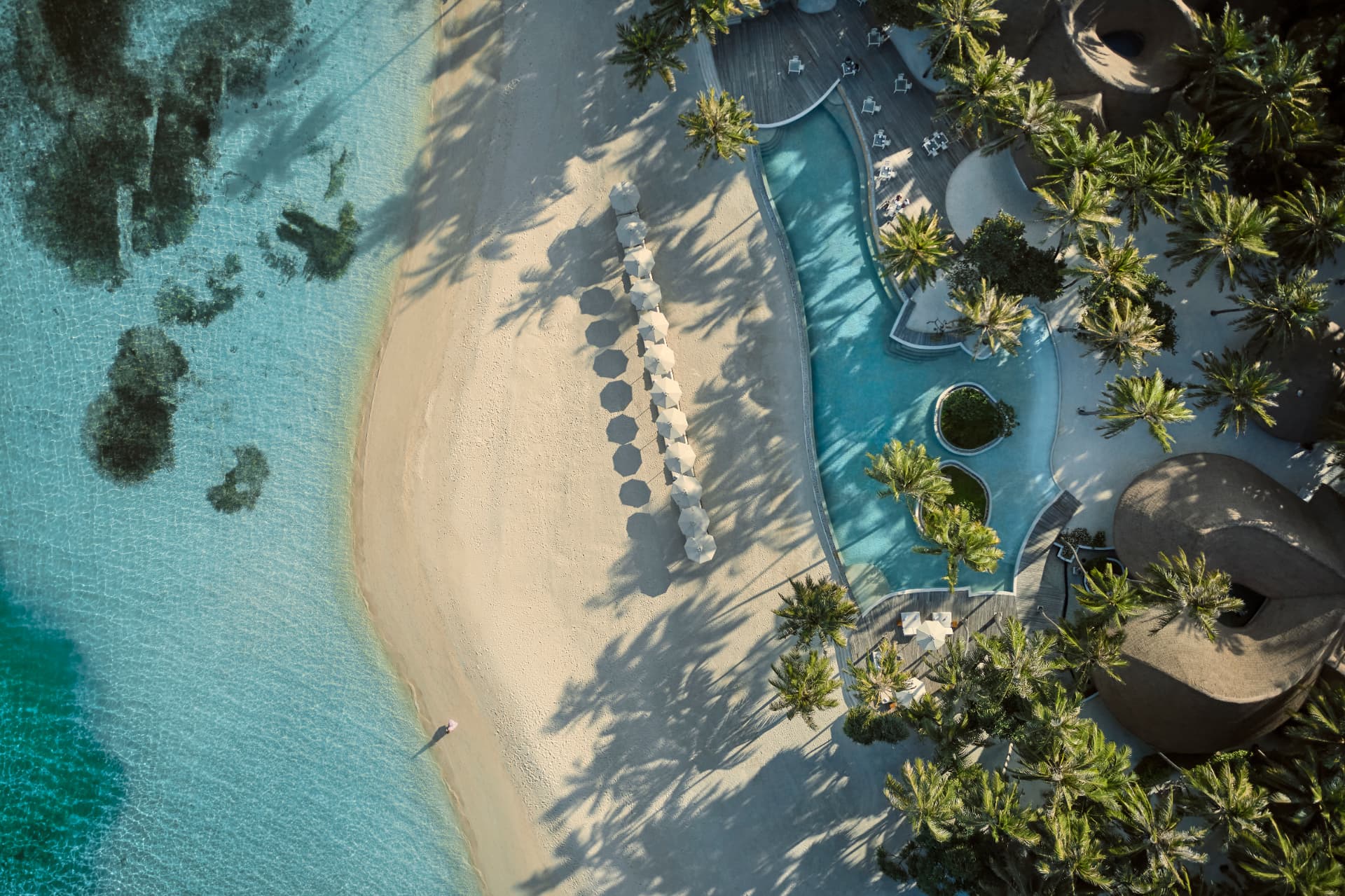 Strand und Pool von oben: helle Schirme, Palmenschatten und klares Lagunenwasser