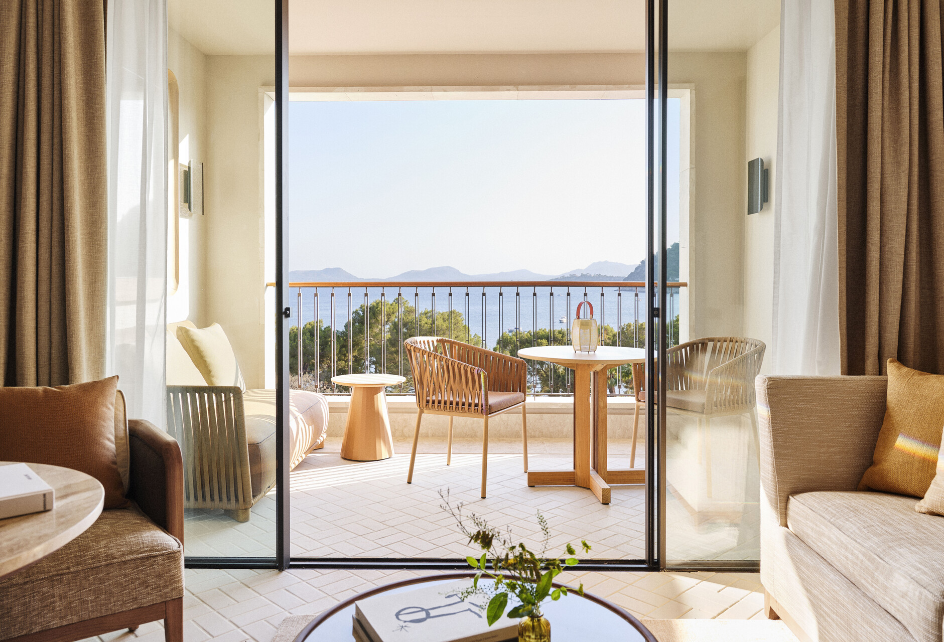 Zimmer mit offener Schiebetür zum Balkon, Korbsesseln und Blick über Pinien auf die Bucht