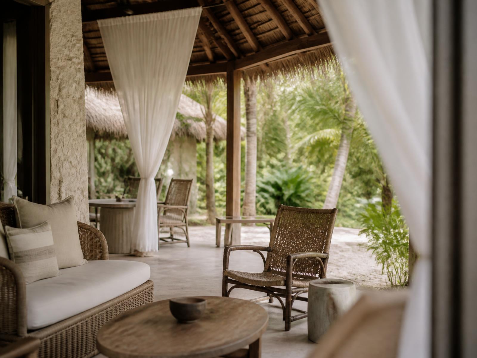 Open-air thatched pavilion with timber beams, coral stone walls, linen curtains and a tropical garden of palms beyond