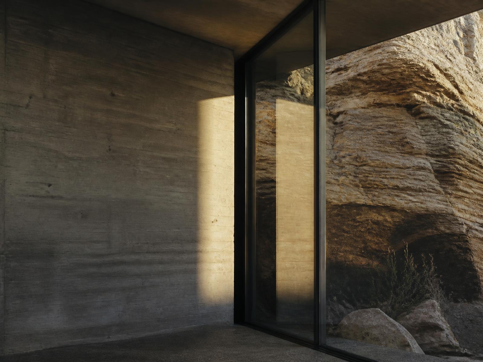 Board-formed concrete wall meeting a natural sandstone rock face behind floor-to-ceiling glass in warm afternoon light