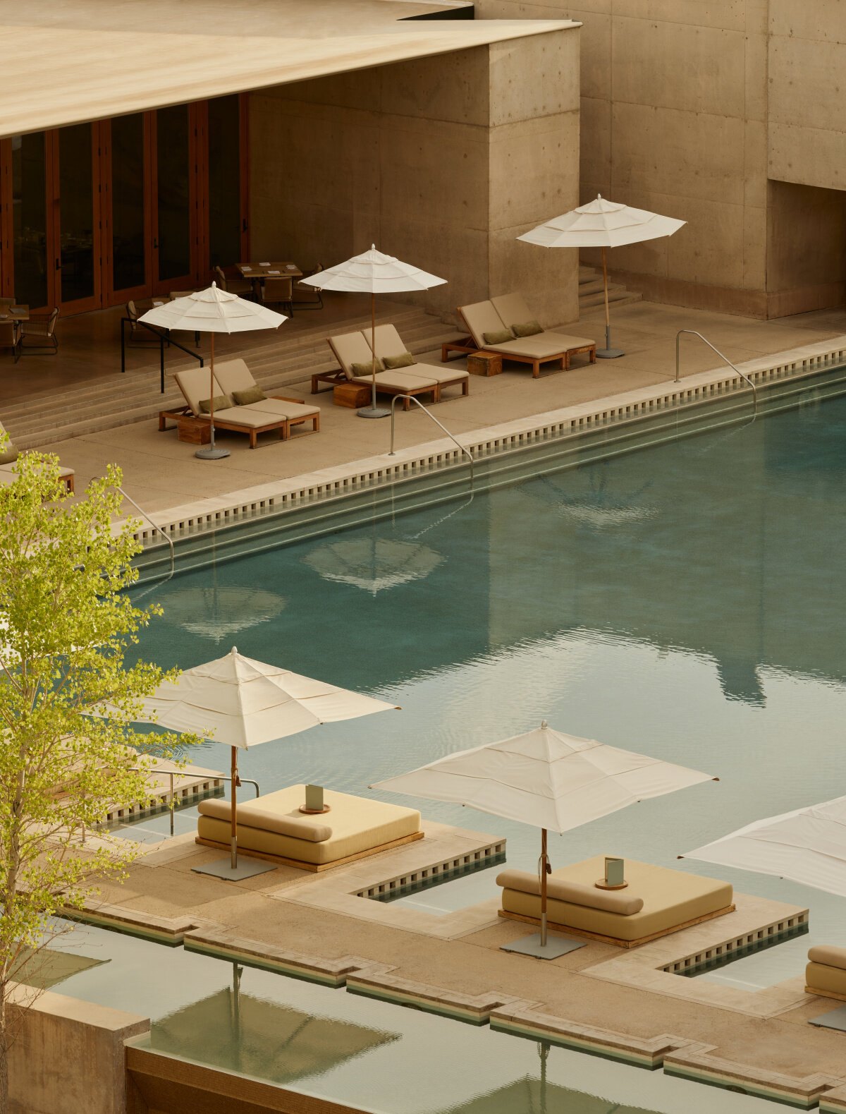 Blick von oben auf die Poolterrasse von Amangiri mit weißen Sonnenschirmen, Teakliegen und der brettergeschalten Betonwand des Hauptpavillons