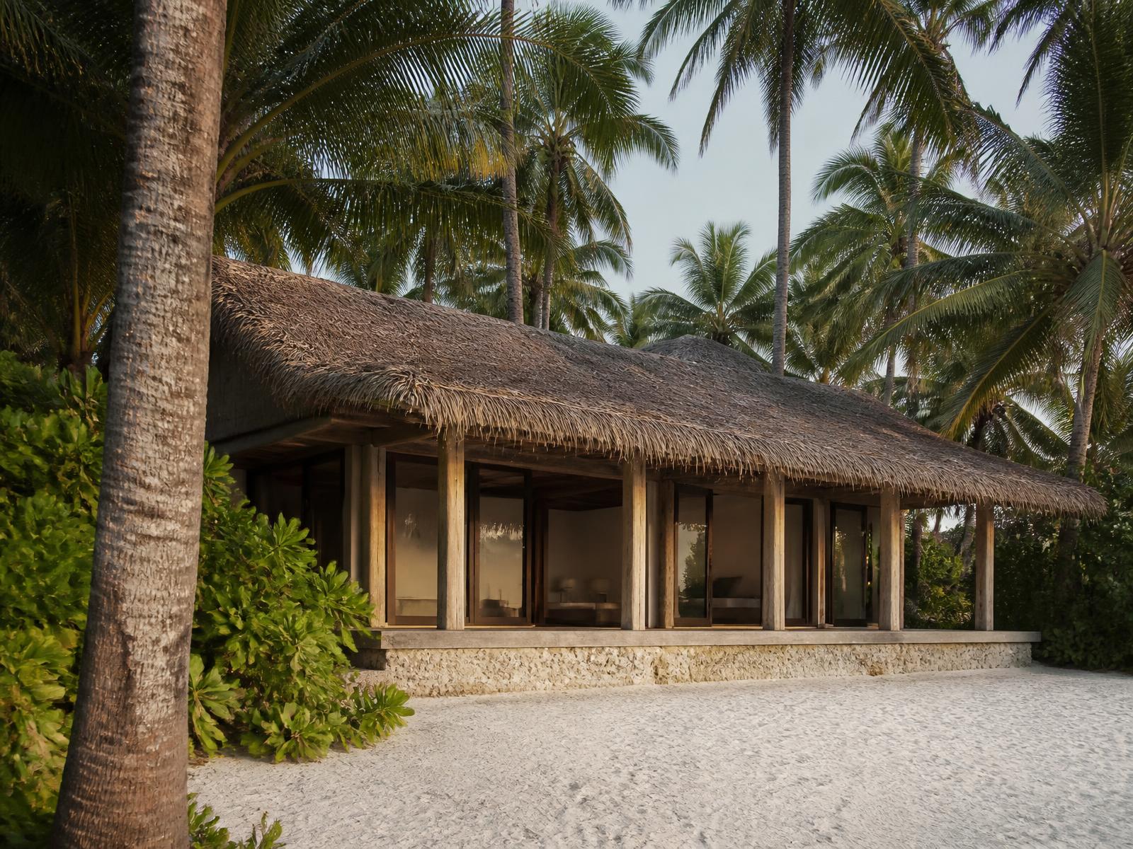 Low thatched-roof timber villa with coral stone base and large glass openings, half hidden among coconut palms on a white sand Polynesian island
