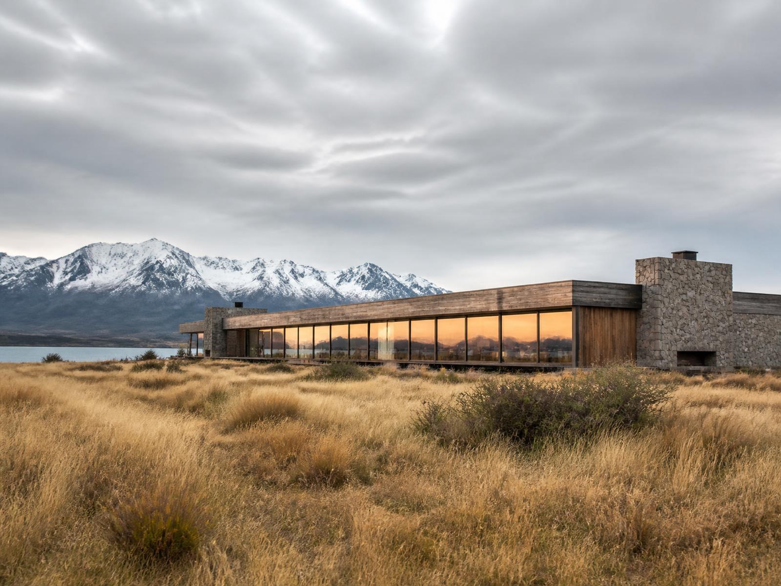Long low minimalist Patagonian lodge of natural timber and local stone with a continuous panoramic window, standing in golden grassland below snow-covered mountains