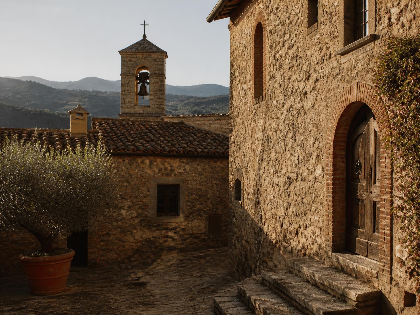 Restored medieval Tuscan borgo with weathered limestone walls, terracotta roofs, an arched wooden door and a stone bell tower in warm afternoon light