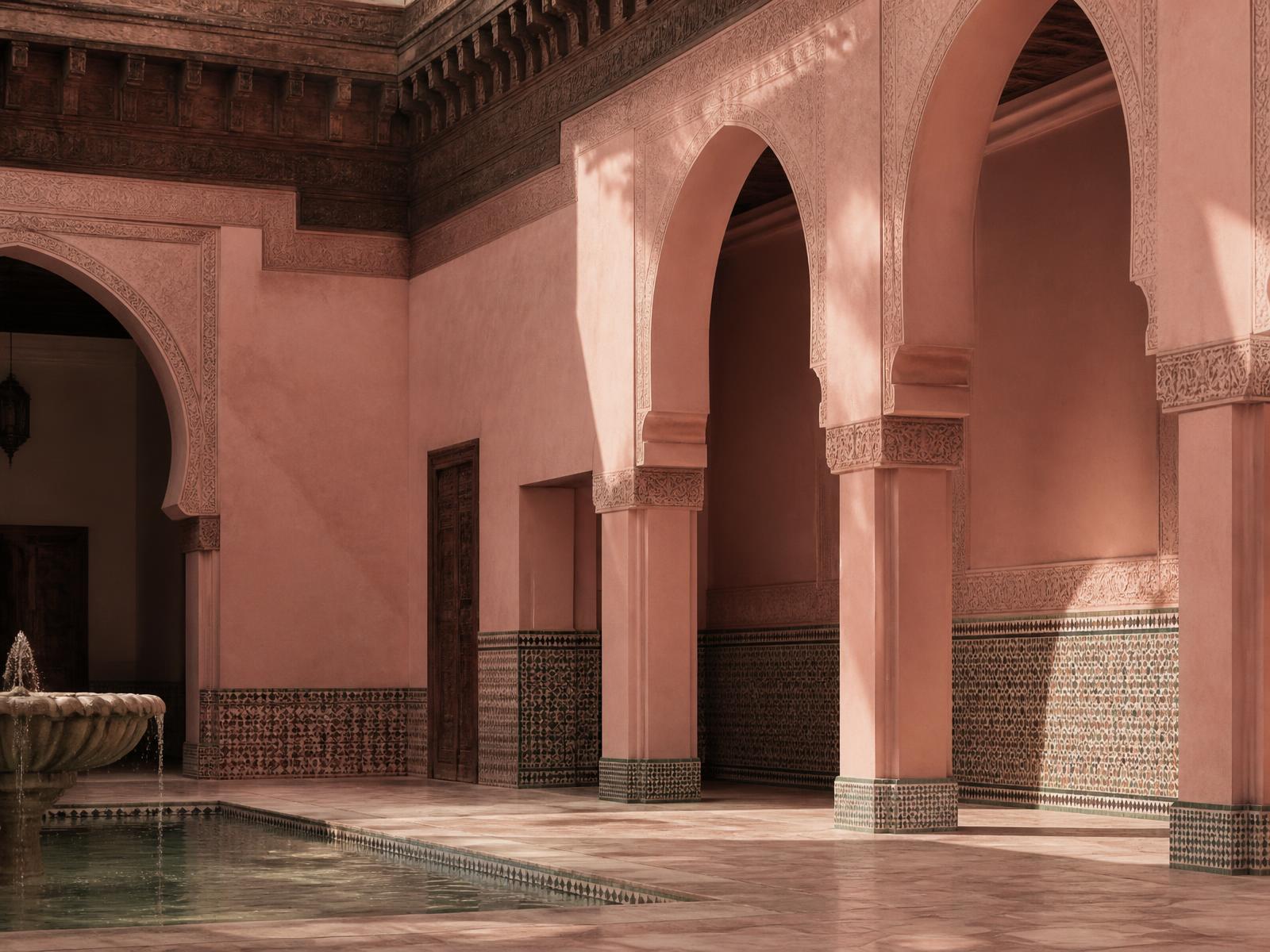 Rose-pink Moorish arches, carved cedar ceiling, zellige tilework and a stone fountain in a quiet Moroccan courtyard in warm natural light
