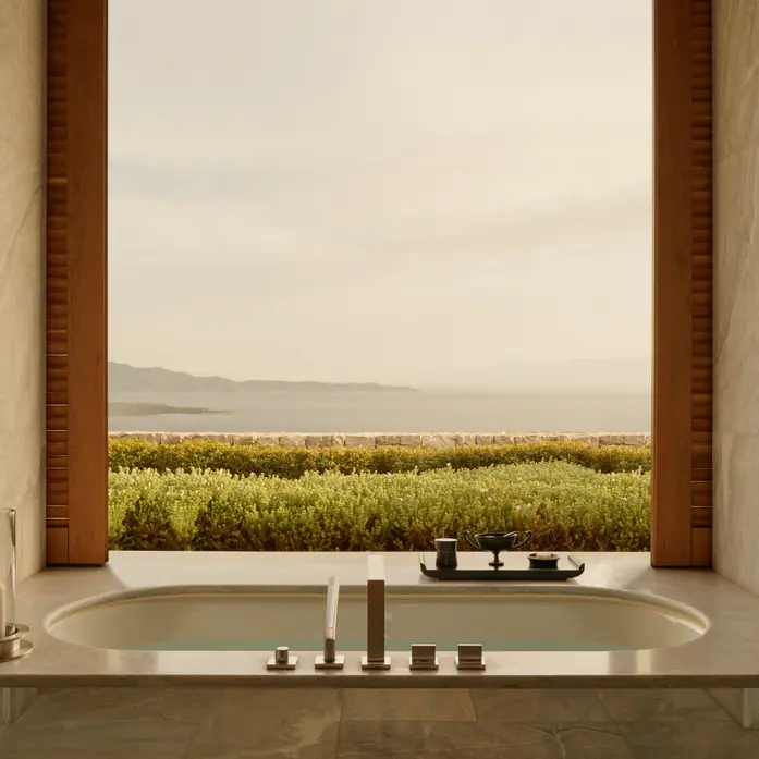 Amanzoe-Bad mit Marmorwanne vor bodentiefer Öffnung, Blick über blühende Hecken auf die diesige Küstenlinie