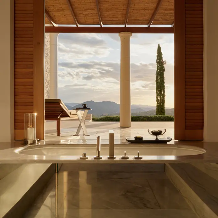 Marmorwanne in einem Amanzoe-Pavillon mit Blick durch geöffnete Holzlamellen auf Säule, Liege, Zypresse und die Berge im Abendlicht