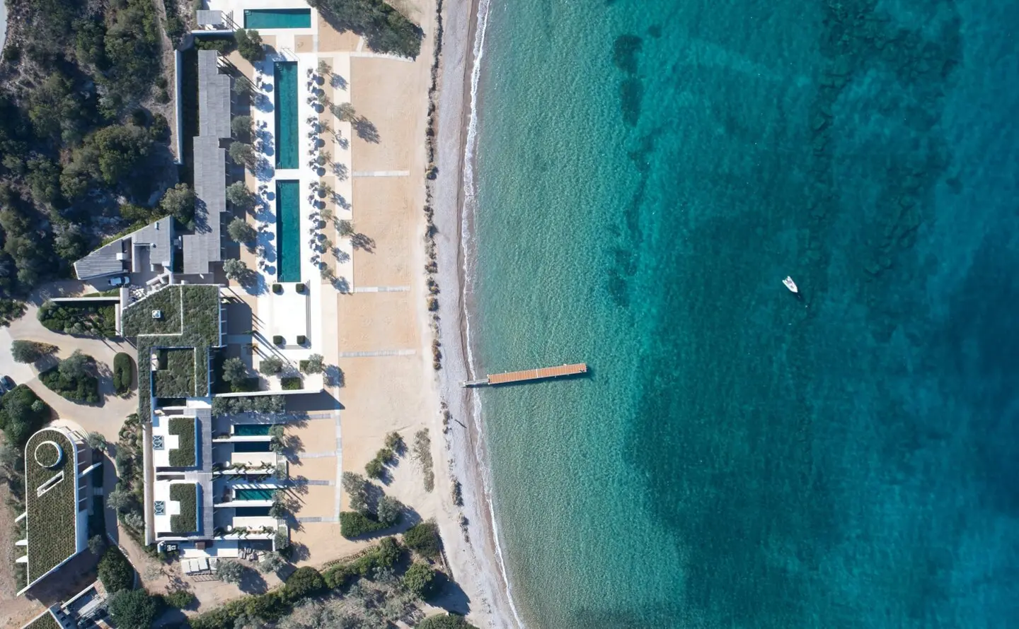 Der Beach Club von Amanzoe aus der Luft: schmale Becken und Cabanas entlang des Sandstrands, ein Holzsteg im türkisfarbenen Wasser