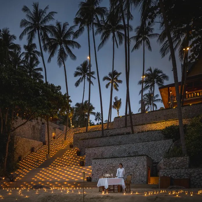 Ein einzelner gedeckter Tisch am Strand bei Nacht, die breite Treppe dahinter mit hunderten Kerzen ausgelegt