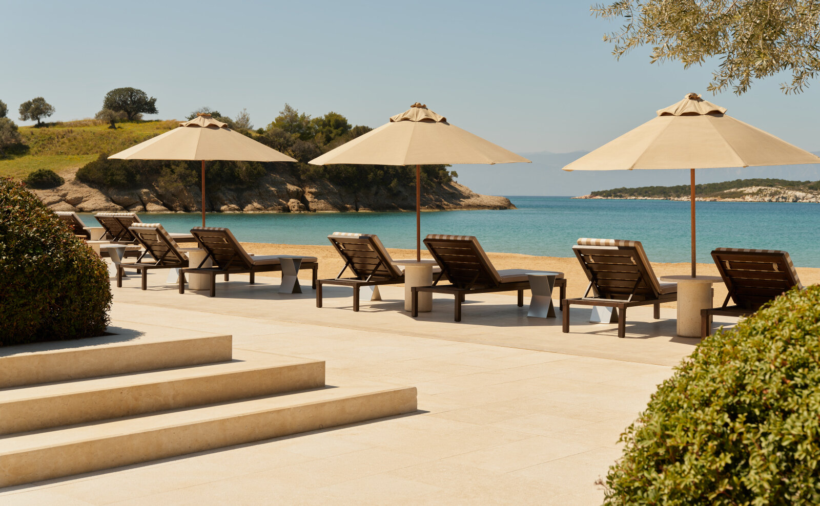 Liegen und Sonnenschirme auf der Steinterrasse des Amanzoe Beach Club, dahinter Sandstrand und die türkisfarbene Bucht