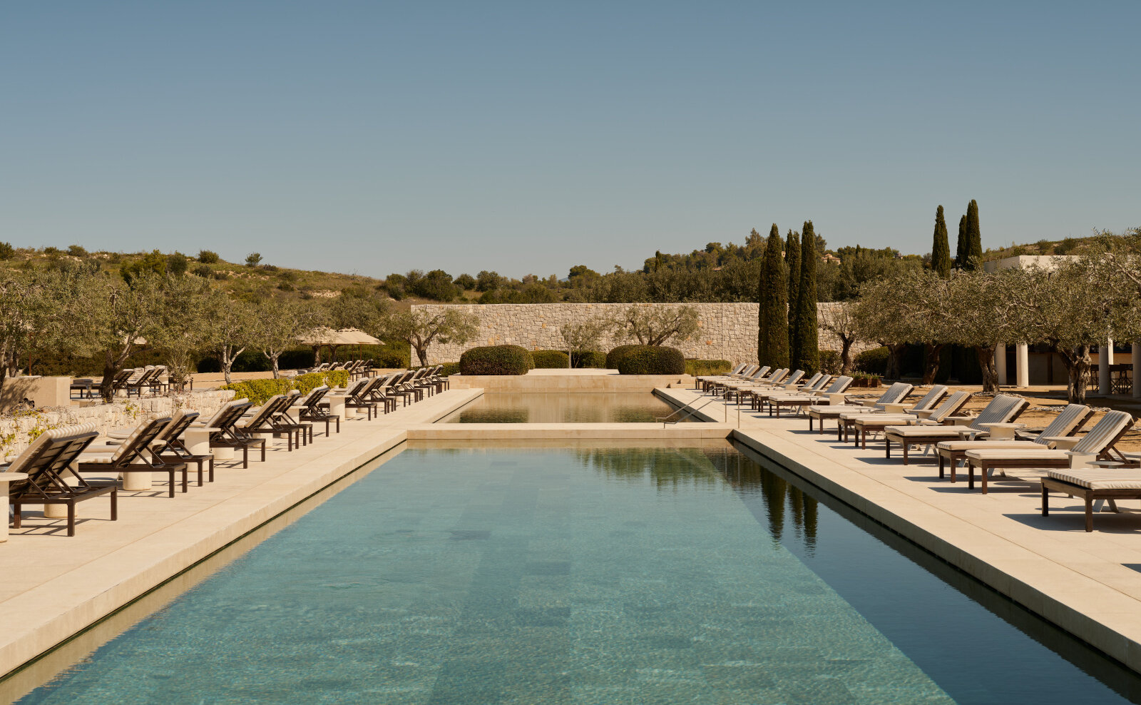 Langes Becken am Beach Club von Amanzoe, gesäumt von Liegen, Olivenbäumen, Zypressen und einer hellen Natursteinmauer