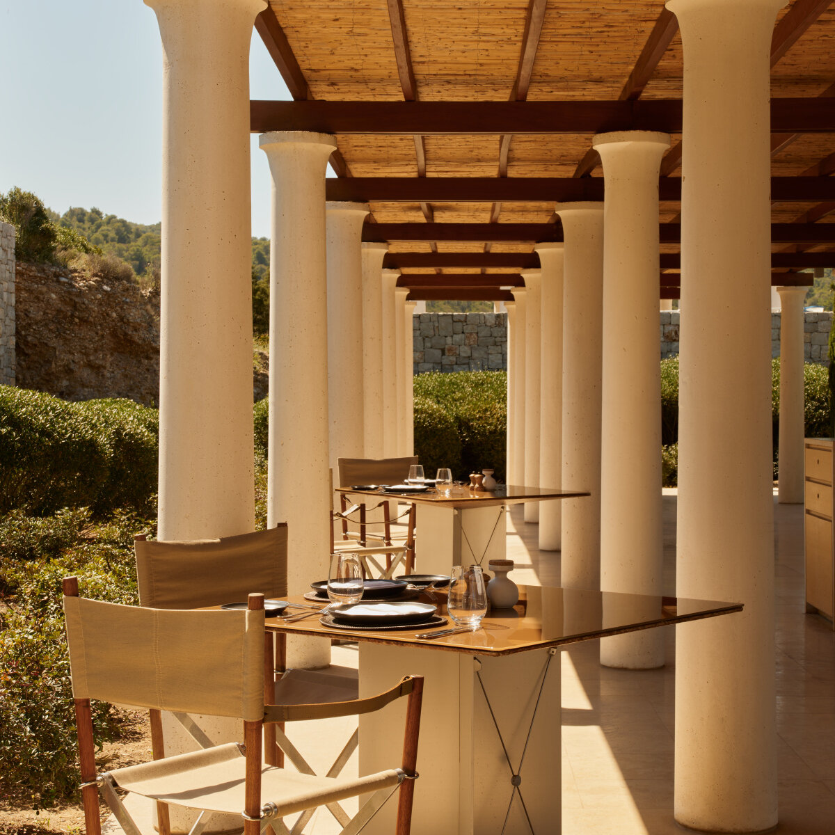 Gedeckte Tische zwischen hellen Säulen im Restaurant des Amanzoe Beach Club, Regiestühle und Schilfdach im Mittagslicht