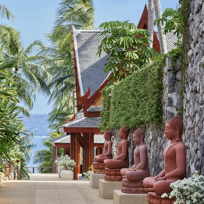 Weg entlang einer Steinmauer mit sitzenden Buddha-Figuren, dahinter ein thailändisches Dach und das Meer