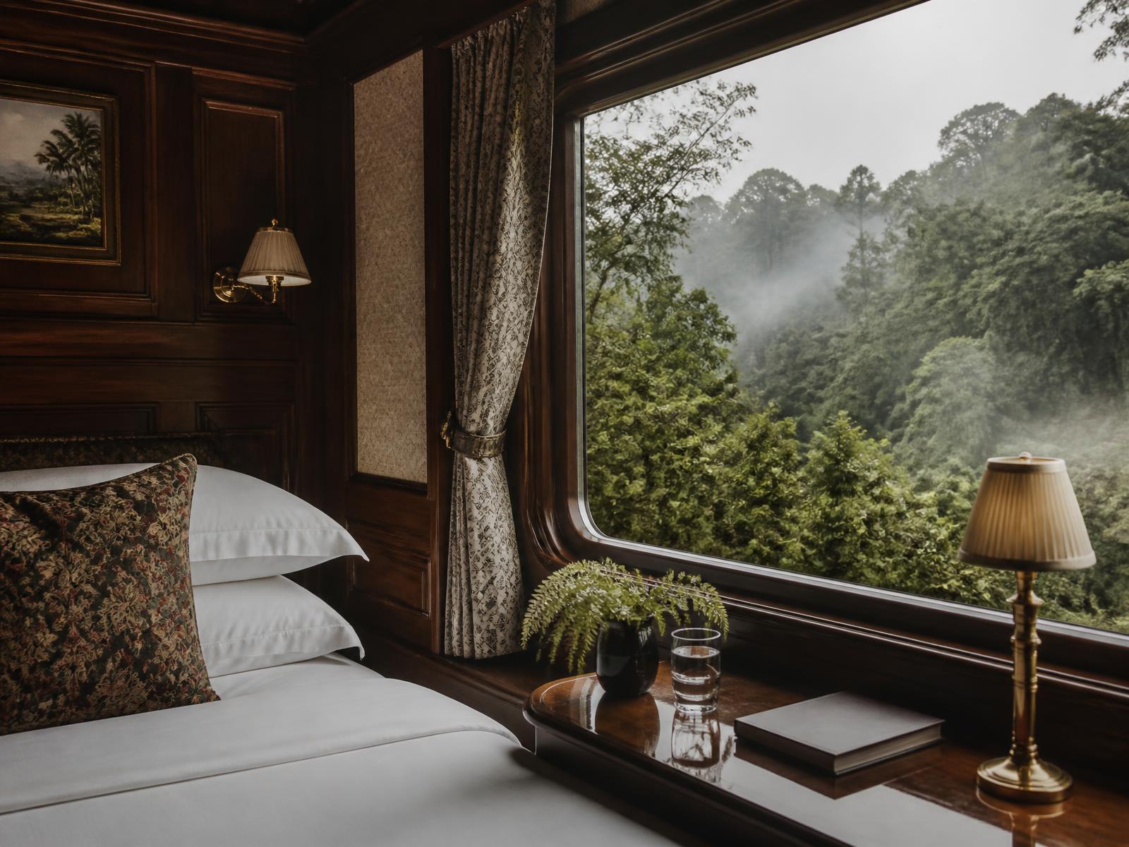 A private cabin aboard a colonial luxury train in Malaysia with timber panelling, white linen, a brass lamp and a large window filled with misty rainforest