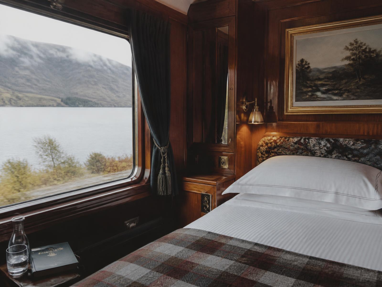 Eine holzgetäfelte Kabine an Bord eines schottischen Luxuszugs mit weißem Leinen, einer gefalteten Tartan-Wolldecke, einer kleinen Messinglampe und einem Fenster mit Blick auf ein nebliges Highland-Loch