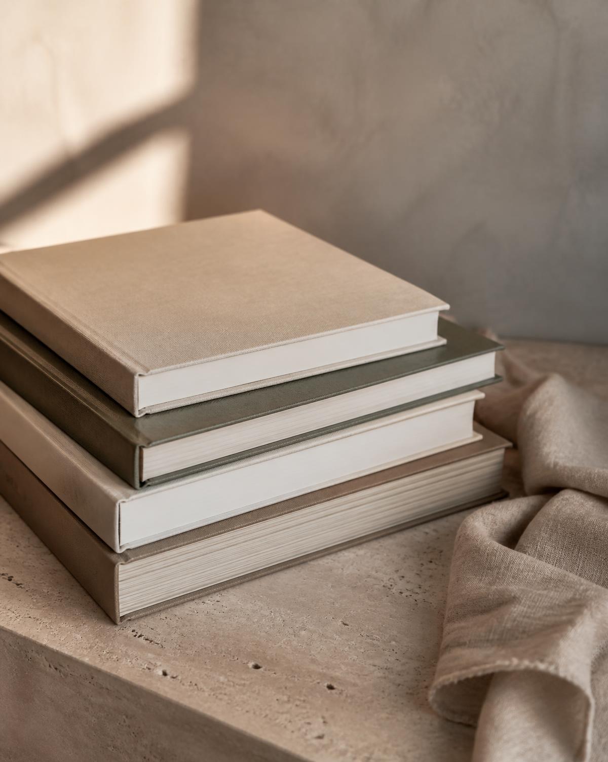 Stack of large neutral-toned coffee table books on a travertine surface beside linen cloth