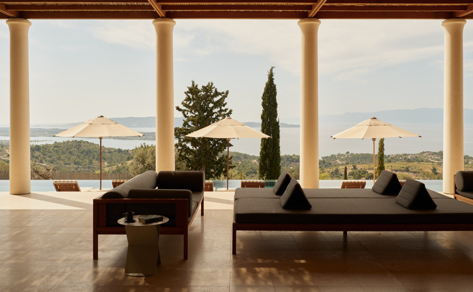 Säulengang einer Amanzoe-Villa mit Daybeds im Schatten, dahinter Infinity-Pool, Zypresse und der Blick auf Porto Heli