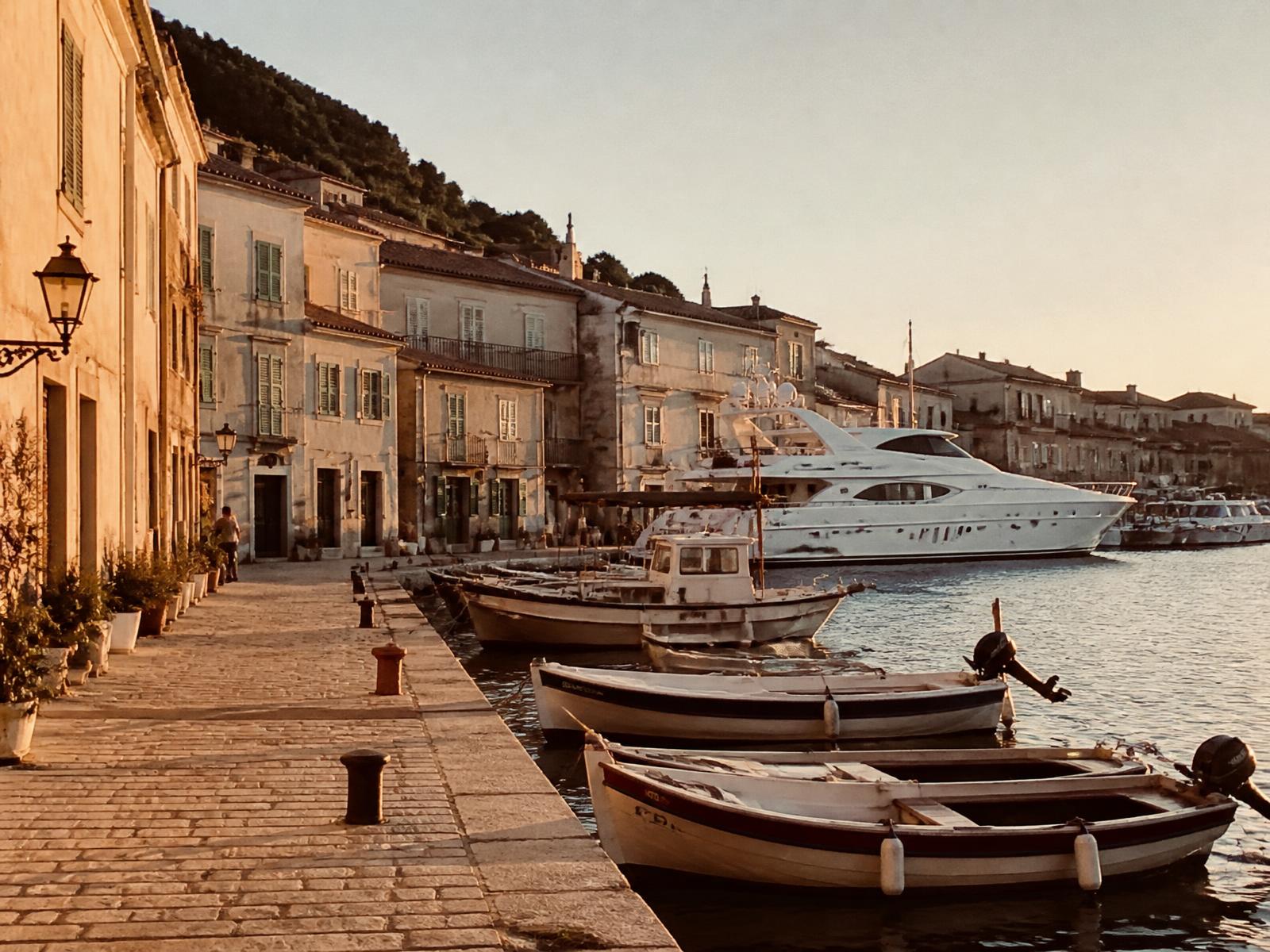 Small Mediterranean fishing harbour at golden hour with pastel stone houses, wooden boats and an elegant white yacht moored at the quay