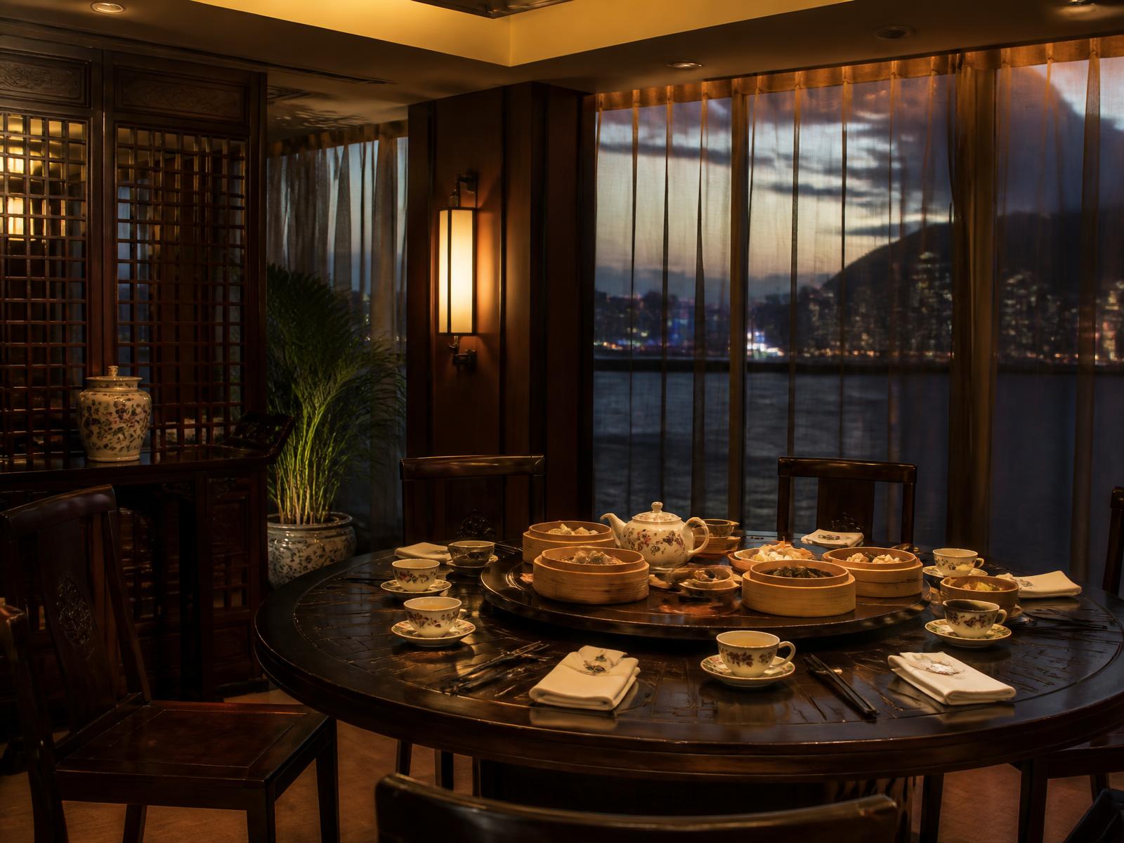 An elegant Cantonese dining room in Hong Kong at dusk with dark lacquer, a round table set with porcelain tea ware and bamboo steamers, harbour lights beyond gauze curtains