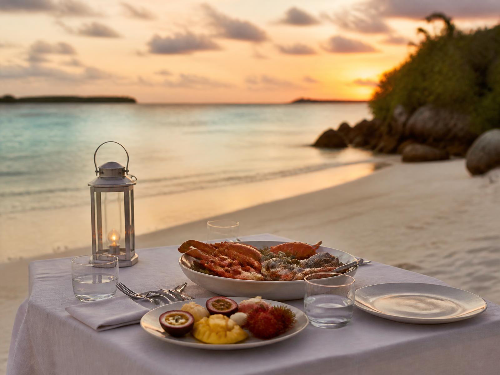 A table set with white linen on white sand at the edge of a turquoise lagoon at sunset, with seafood, tropical fruit and a lantern