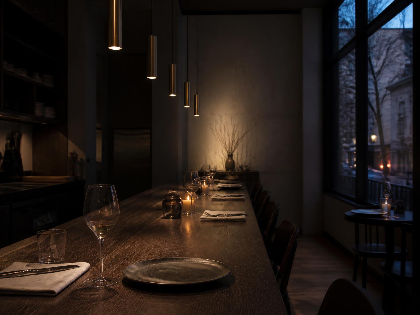 Intimate dark oak dining counter with bronze pendant lights, candlelight, simple ceramic plates and a single glass of wine at winter dusk
