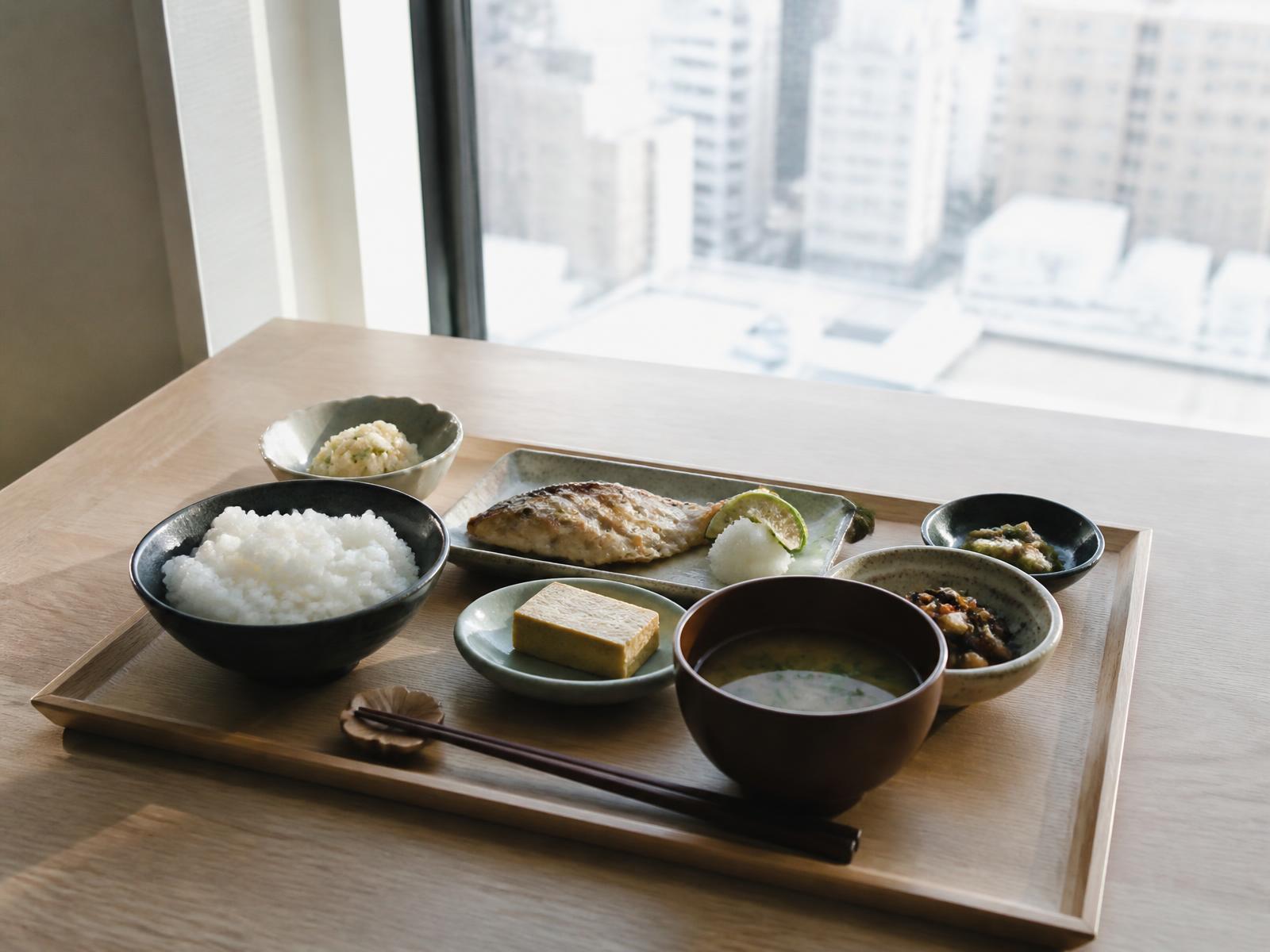 Japanese breakfast tray of small ceramic dishes, grilled fish, rice and miso on pale wood beside a tall window over the city