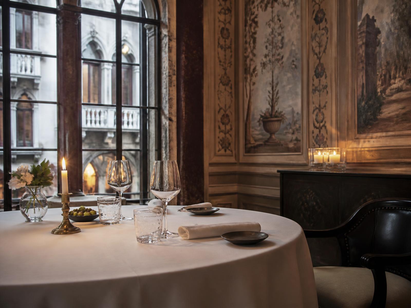 Single table set with white linen and glassware in a frescoed Venetian dining room lit by candles