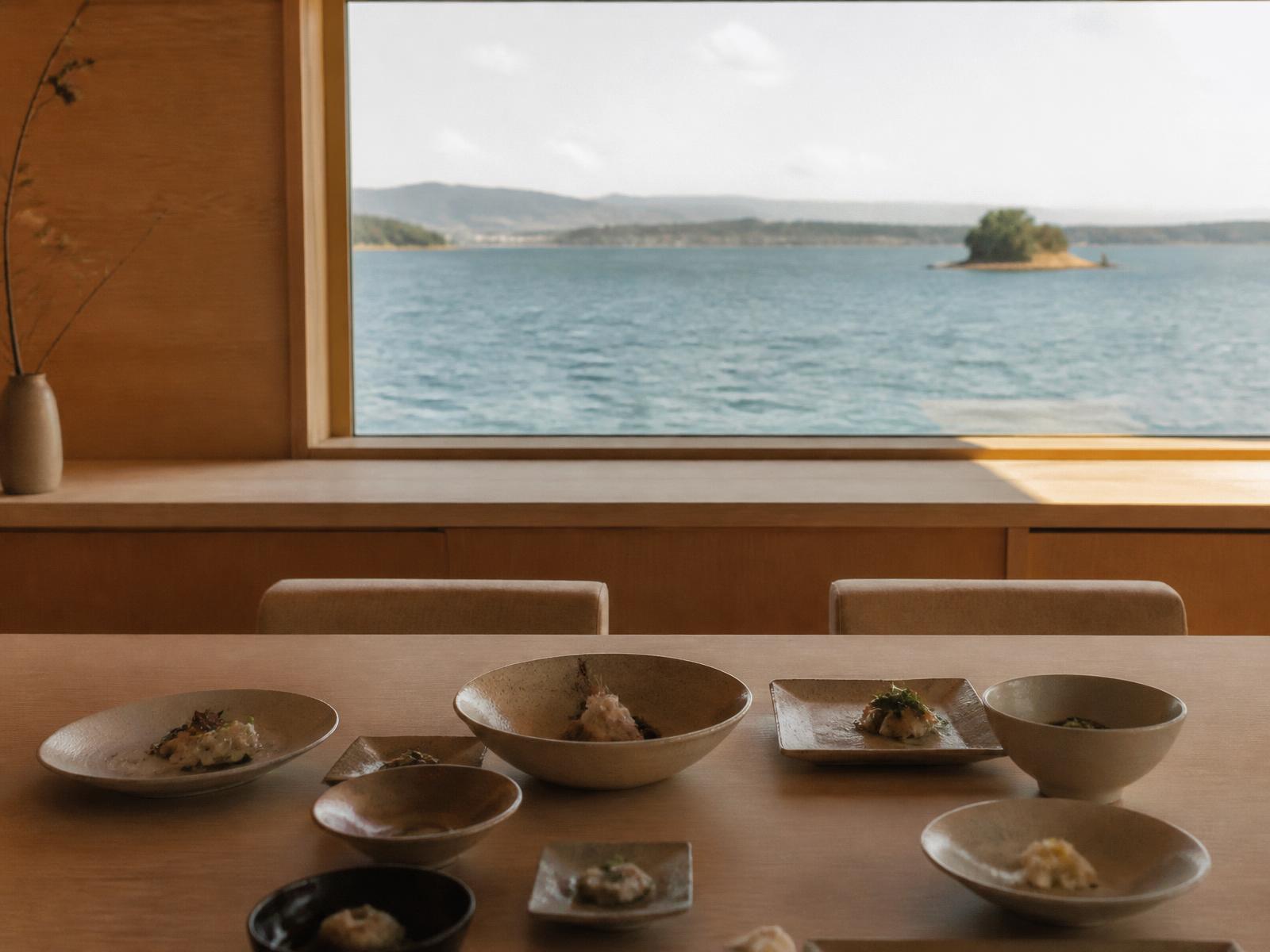 Low hinoki dining counter set with ceramic bowls and small plates, wide window onto a calm sea
