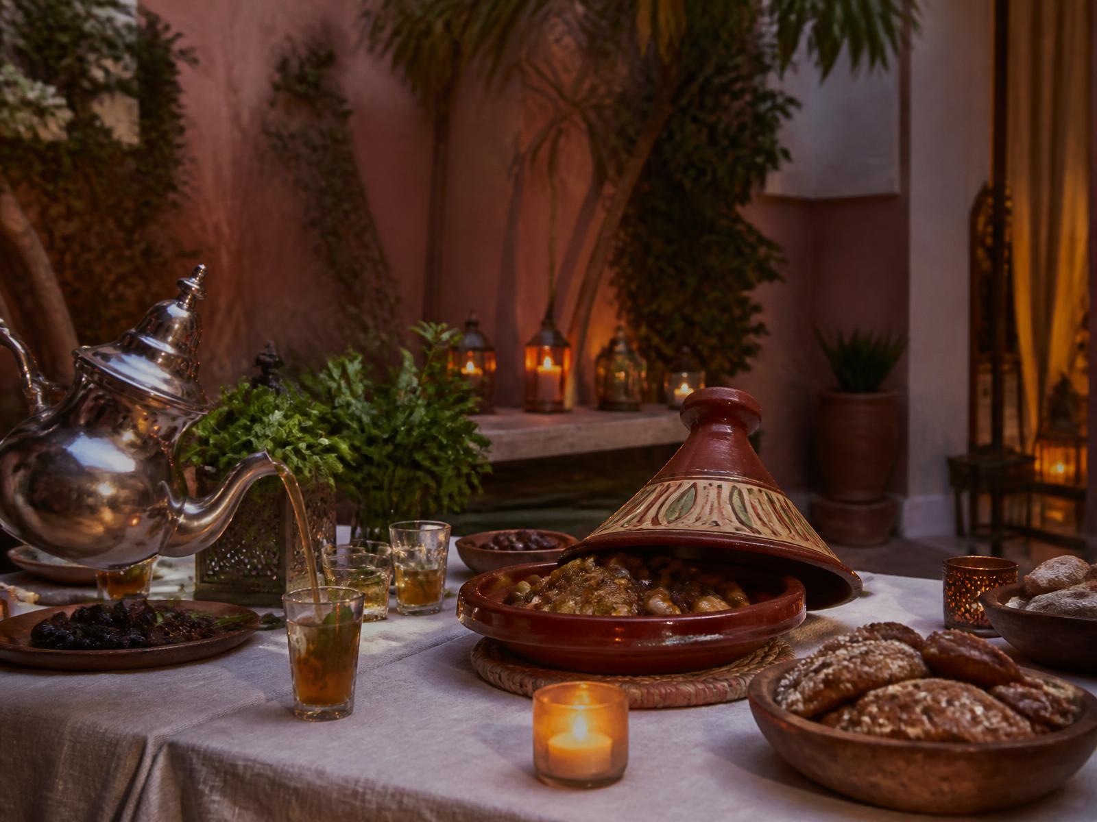 A candlelit table in a rose-walled Moroccan courtyard at dusk with a traditional tagine, mint tea poured from a silver pot, garden herbs and Moroccan pastries