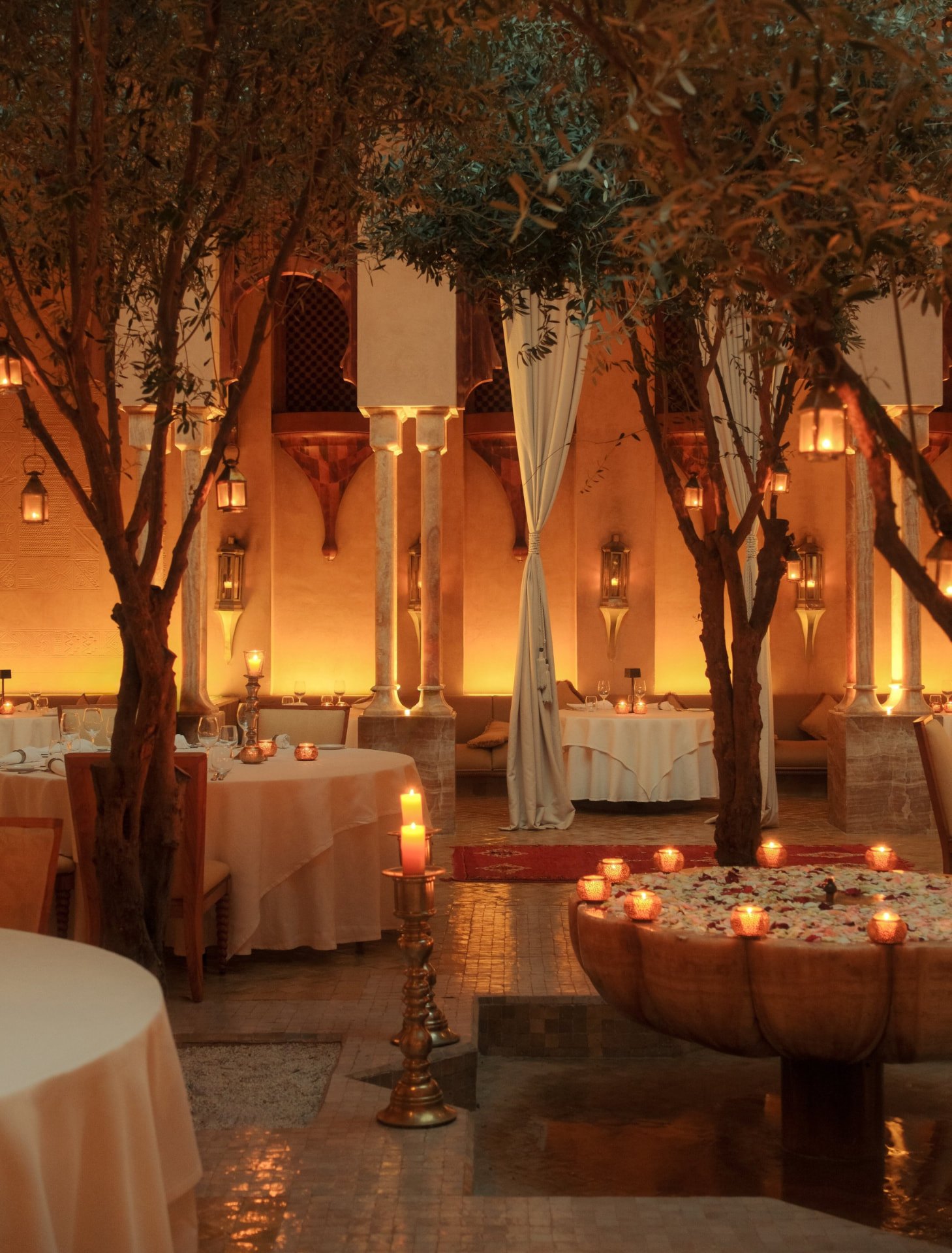 Das marokkanische Restaurant von Amanjena bei Nacht: Olivenbäume, Laternen, Messingleuchter und ein mit Rosenblättern bedecktes Wasserbecken