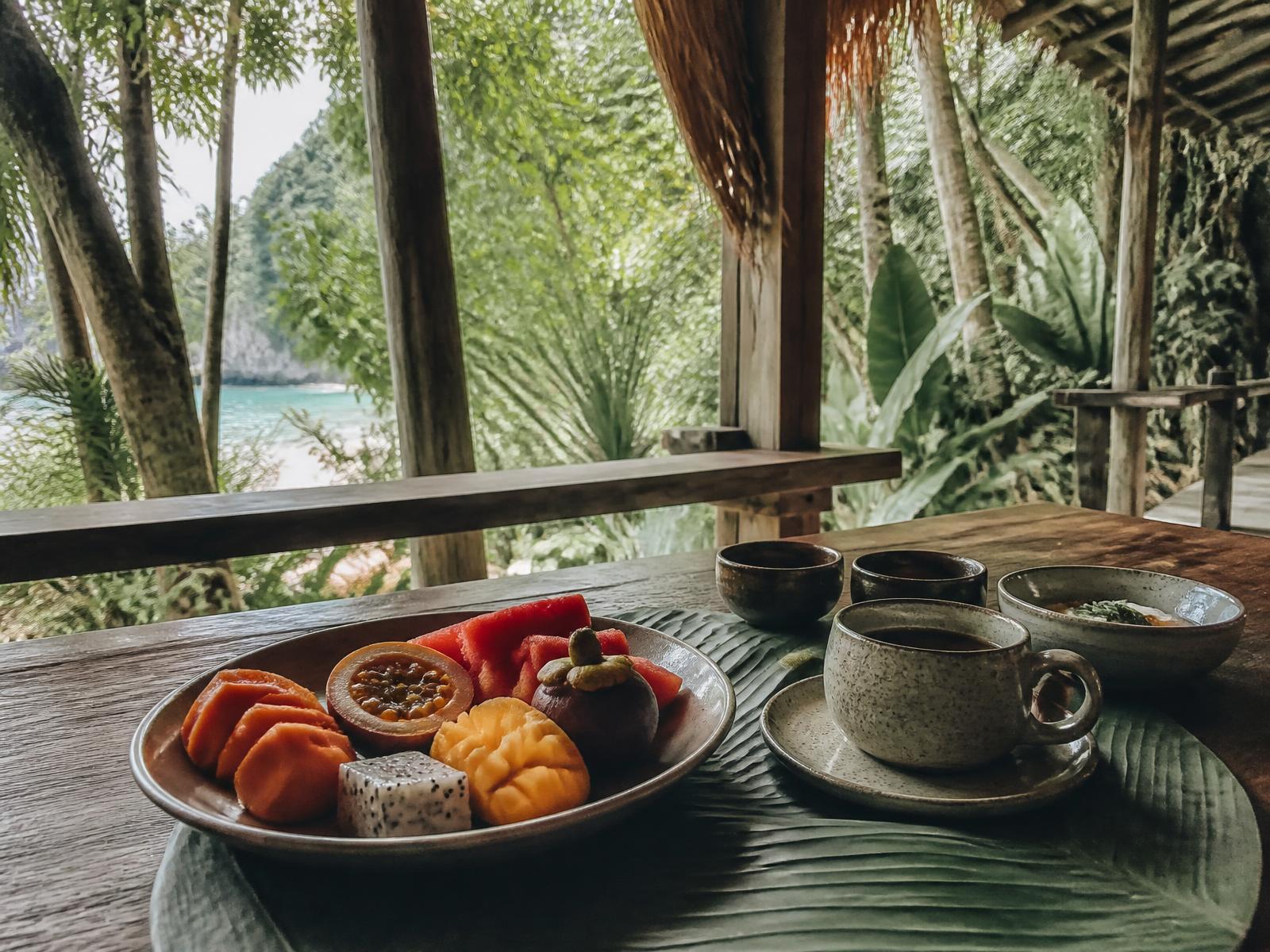Slow tropical breakfast on a timber table in an open-air rainforest pavilion with fresh fruit, local coffee and a sliver of emerald sea beyond