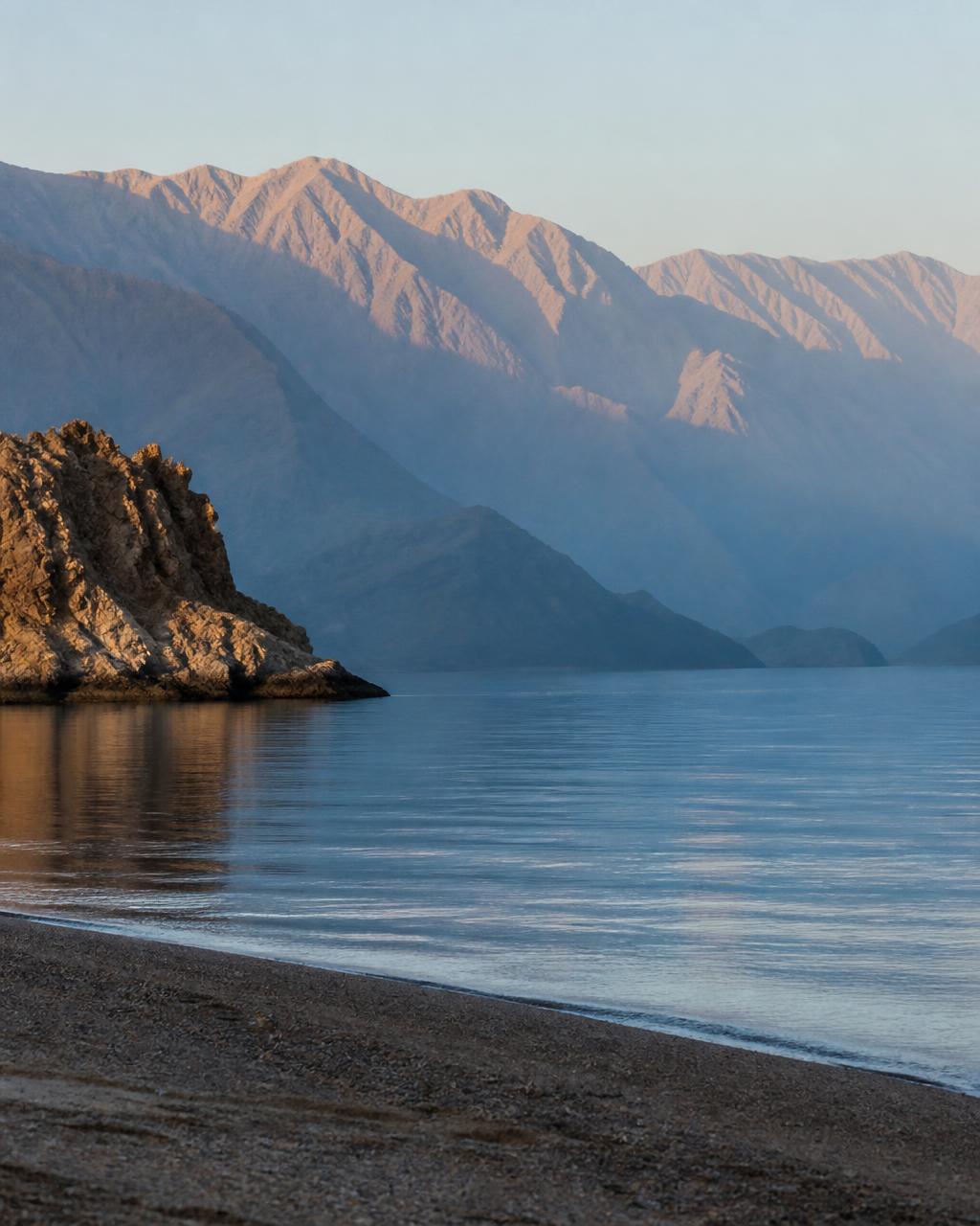 First light on the bare ridges of the Hajar Mountains above a still bay, layered blue and ochre silhouettes with an empty beach in the foreground