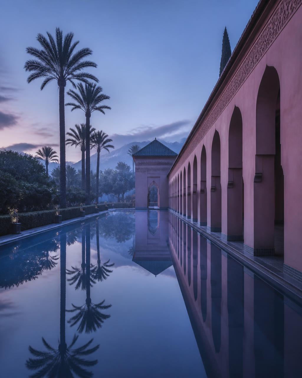 A long still reflecting pool at dawn in a Marrakech garden mirroring a rose-coloured arcade, date palms and the Atlas Mountains